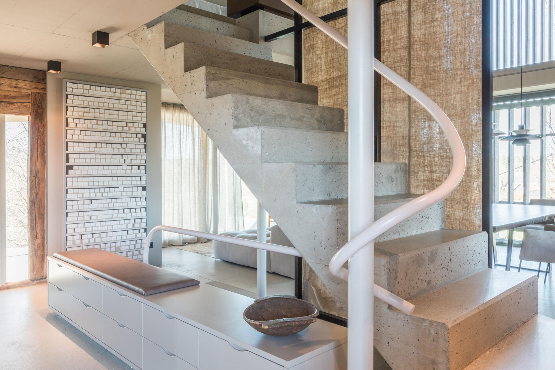 A sculptural concrete staircase in an Austrian farmhouse renovation features angular steps, a pale pink curved handrail, and a custom storage bench with upholstered seating.