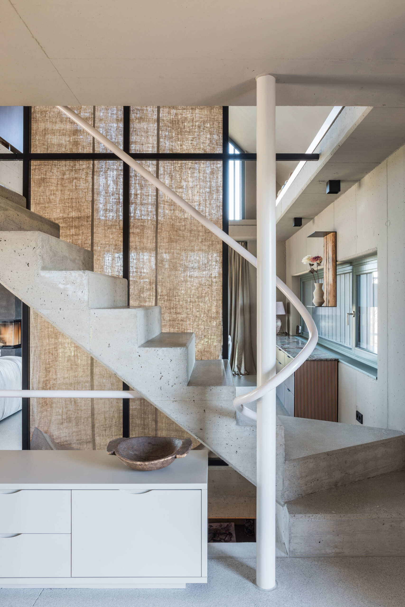 A sculptural concrete staircase in an Austrian farmhouse renovation features angular steps, a pale pink curved handrail, and a custom storage bench with upholstered seating.