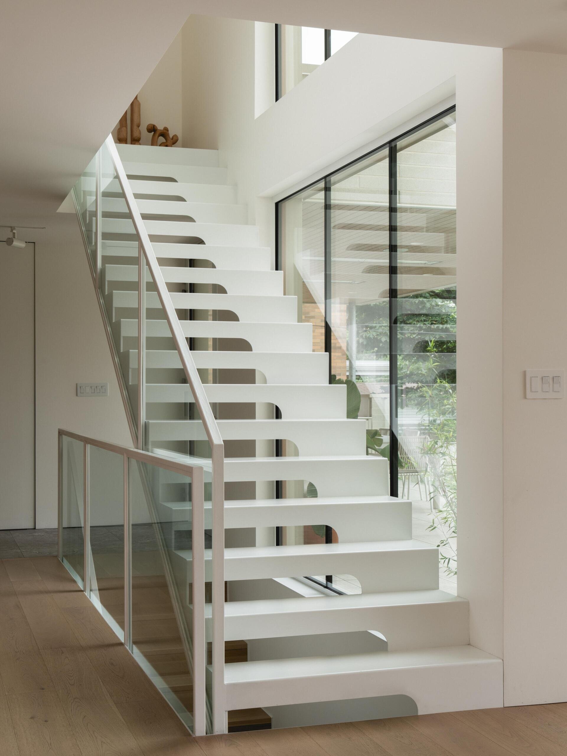 See the floating white steel staircase inside The Lens by MU Architecture in Montreal. Open risers, glass railings, tall windows, and minimalist upper hallways create a bright architectural feature spanning multiple floors.
