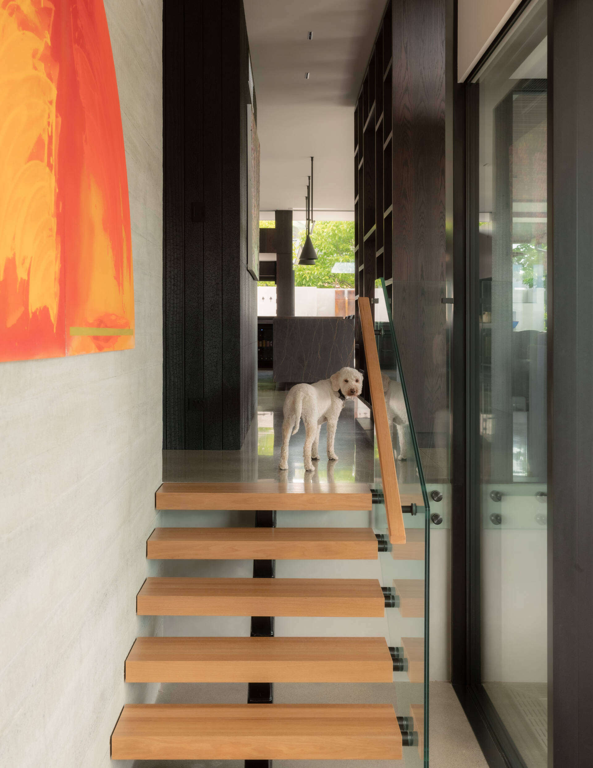 Modern floating wood stairs with open treads, a glass balustrade, and a minimalist handrail, complemented by a colorful abstract painting on a concrete wall.