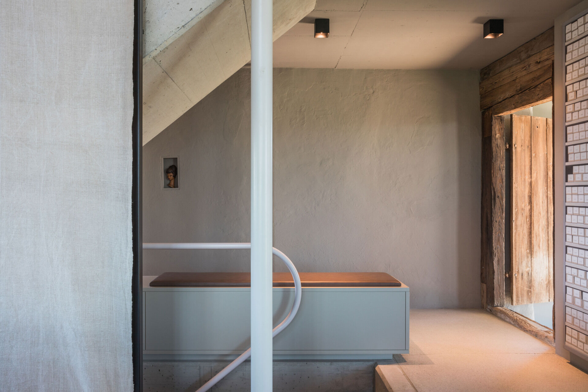 A sculptural concrete staircase in an Austrian farmhouse renovation features angular steps, a pale pink curved handrail, and a custom storage bench with upholstered seating.