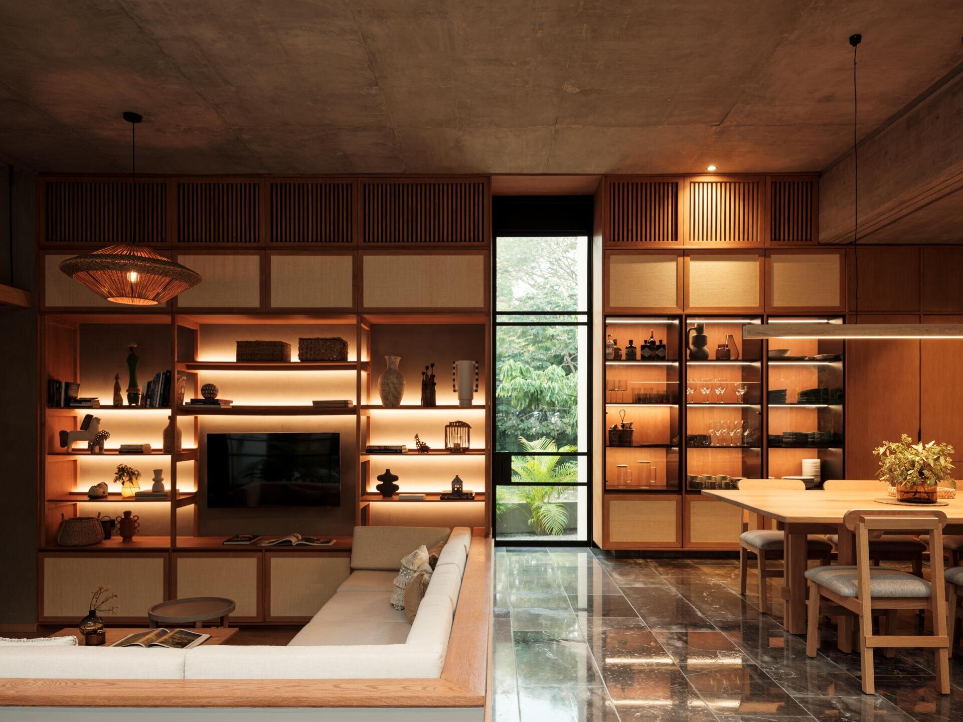 Discover the sunken living room at Casa Akina in Mexico, featuring a built-in L-shaped sofa, custom wood cabinetry, illuminated shelving, woven furniture, and a warm, modern interior design.