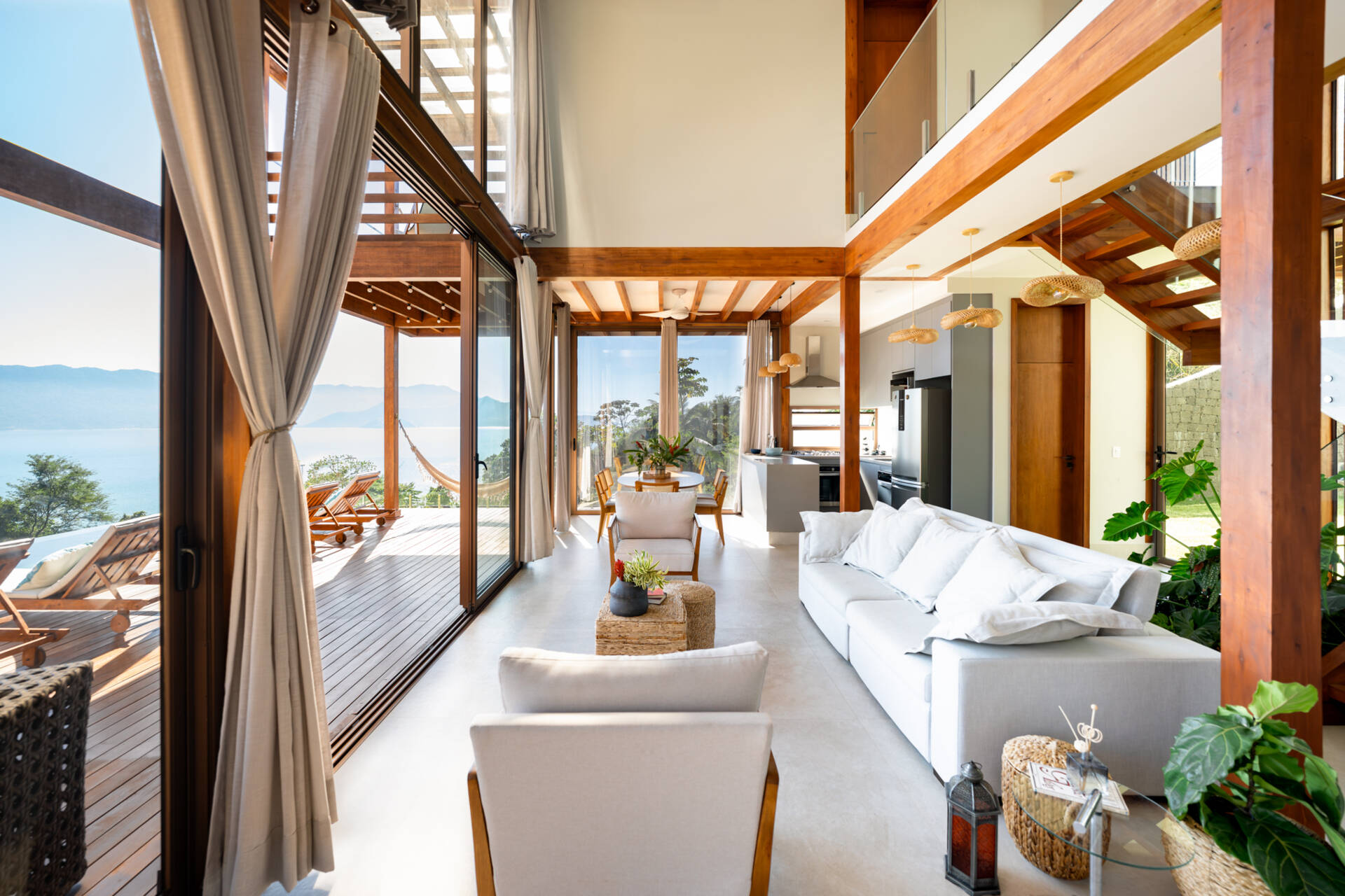 Inside Casa Ponta da Sela in Ilhabela, exposed timber framing, double-height space and large sliding glass doors define an open living, dining and kitchen area with wide views toward the ocean and tropical landscape.