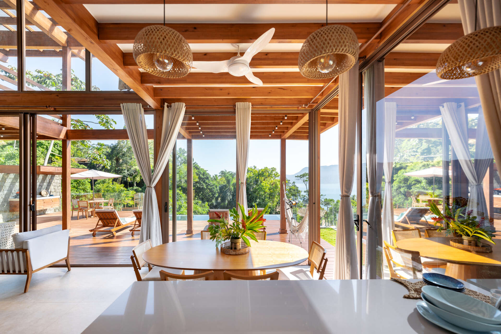 Inside Casa Ponta da Sela in Ilhabela, exposed timber framing, double-height space and large sliding glass doors define an open living, dining and kitchen area with wide views toward the ocean and tropical landscape.