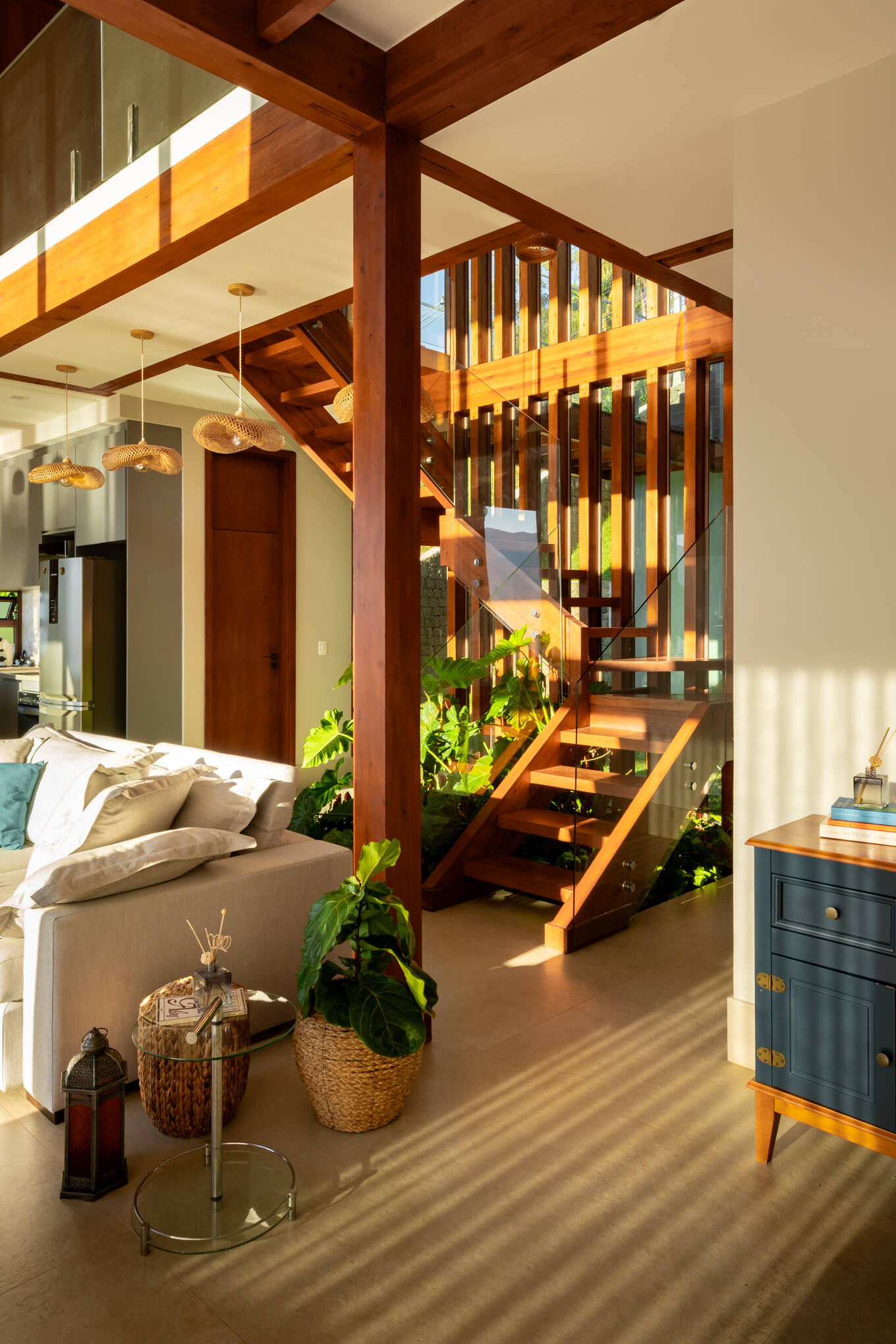 A timber staircase rises through the double-height center of Casa Ponta da Sela, with open treads, glass balustrades and tropical planting tucked beneath the structure to bring greenery into the heart of the home.