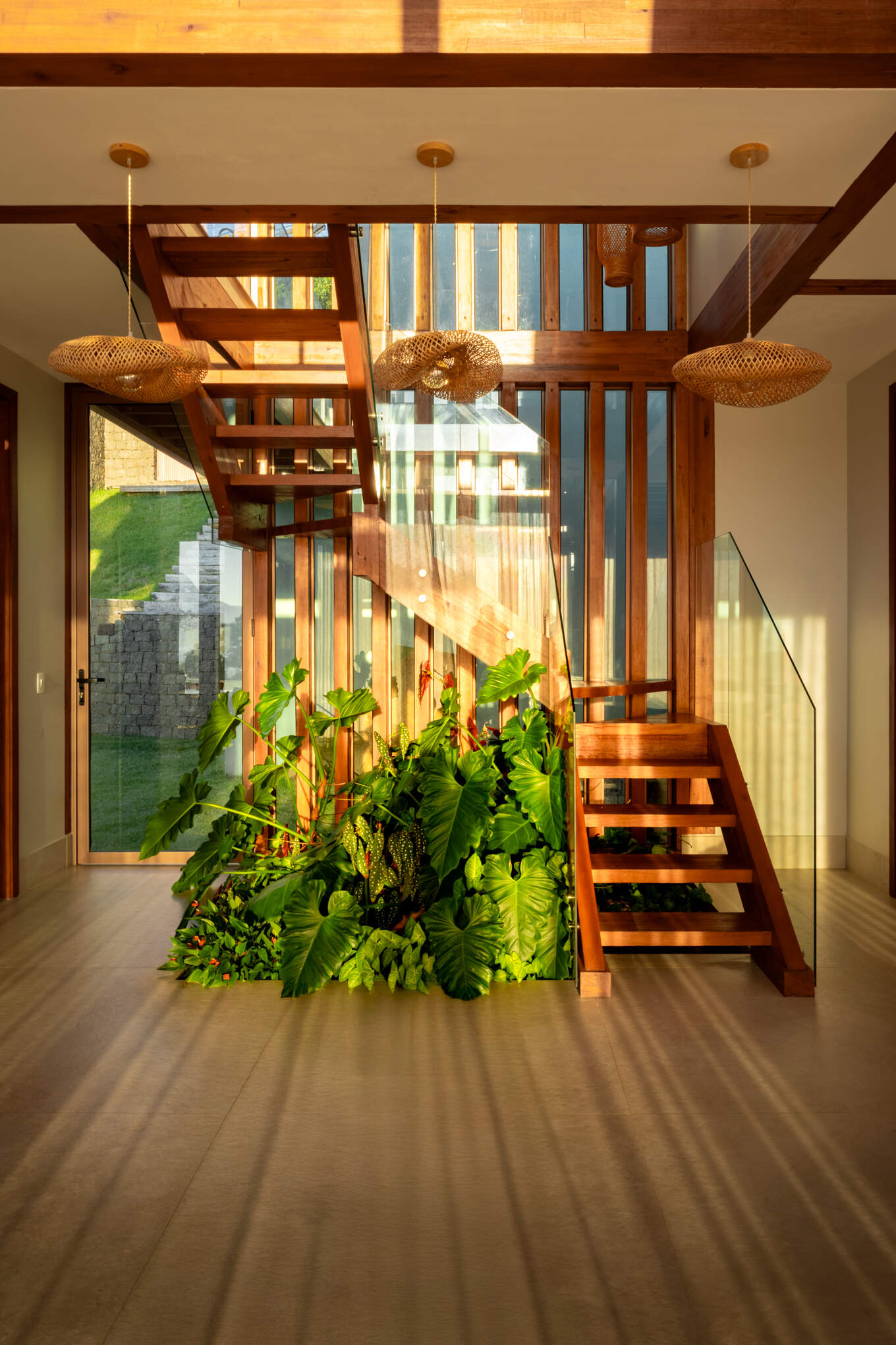 A timber staircase rises through the double-height center of Casa Ponta da Sela, with open treads, glass balustrades and tropical planting tucked beneath the structure to bring greenery into the heart of the home.