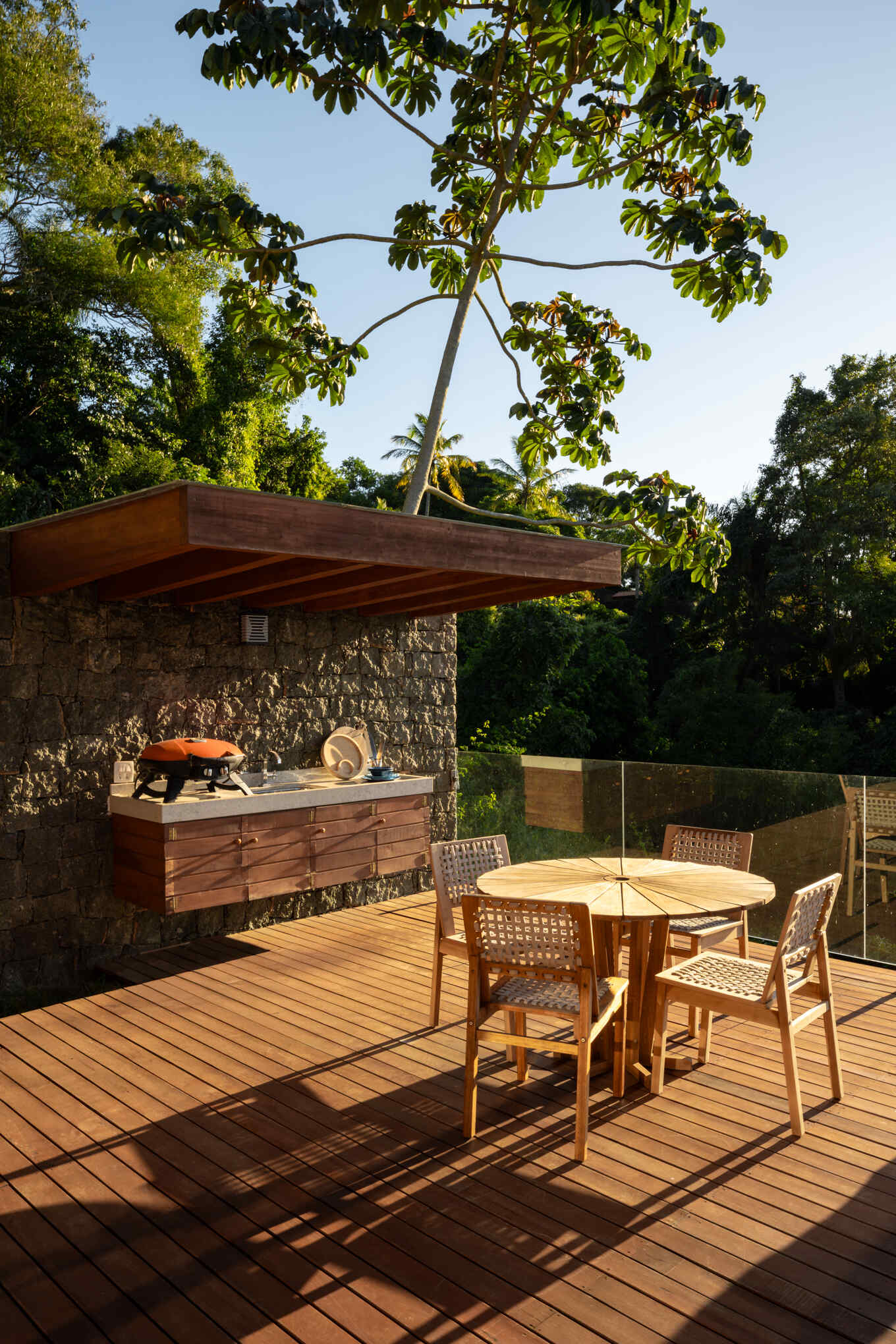 The outdoor kitchen at Casa Ponta da Sela pairs timber cabinetry and decking with a dark stone wall and simple dining area, creating a relaxed entertaining space beside the pool and surrounding tropical landscape.