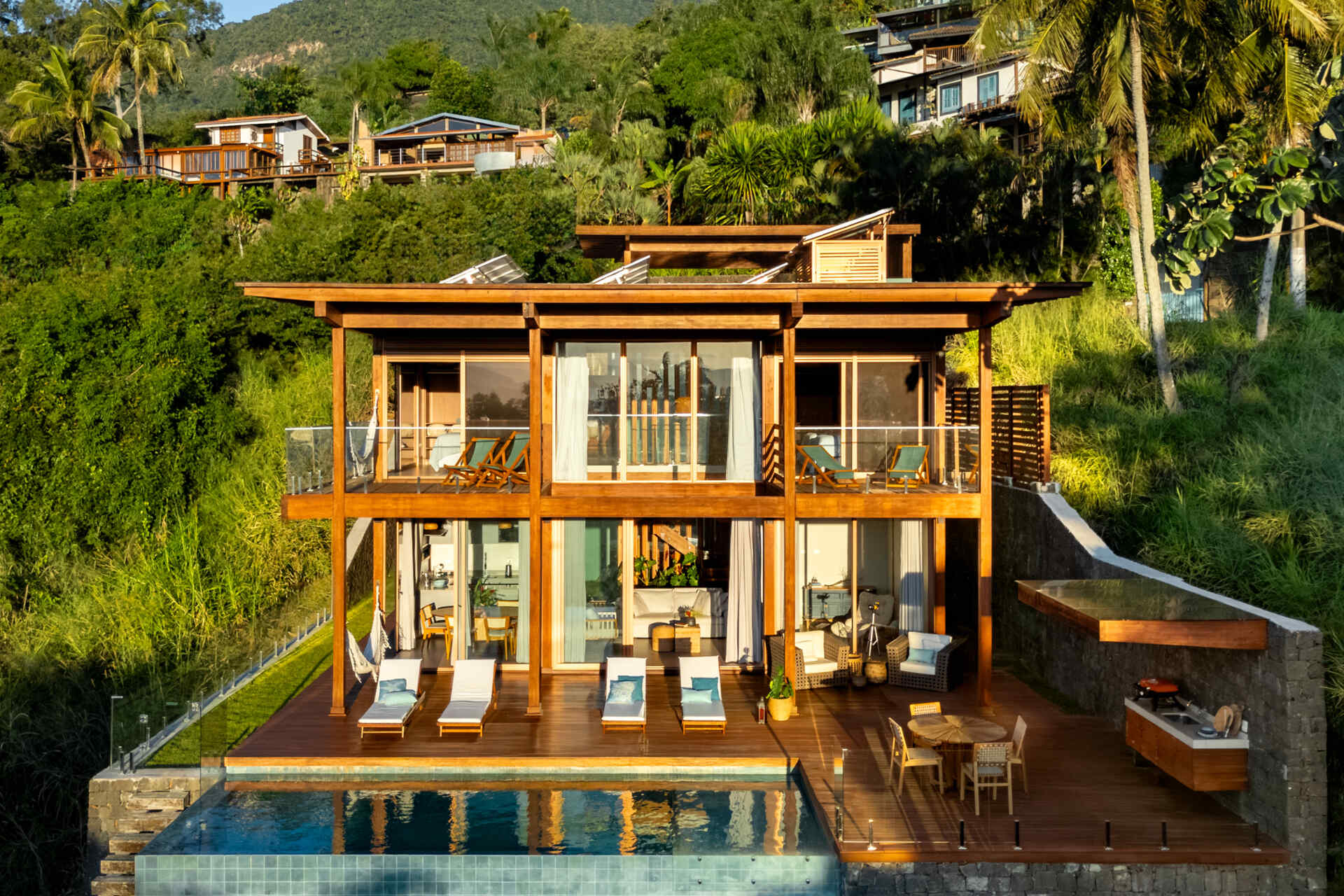 Casa Ponta da Sela in Ilhabela, Brazil, uses prefabricated timber construction, stone retaining walls, glass and timber decks to step down a steep tropical site. Existing rocks, a flat roof with solar systems and an elevated pool become part of the architecture.