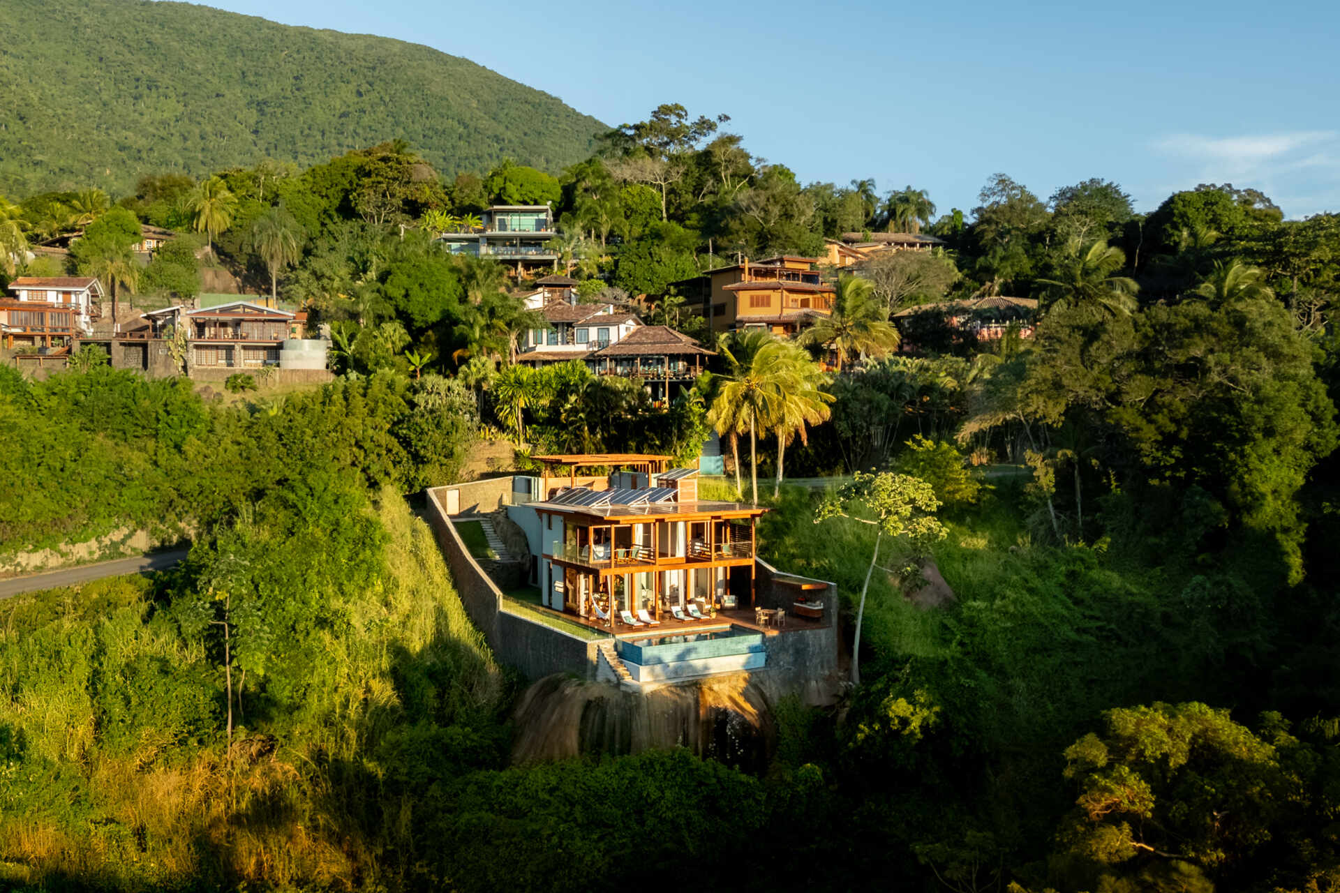 Casa Ponta da Sela in Ilhabela, Brazil, uses prefabricated timber construction, stone retaining walls, glass and timber decks to step down a steep tropical site. Existing rocks, a flat roof with solar systems and an elevated pool become part of the architecture.