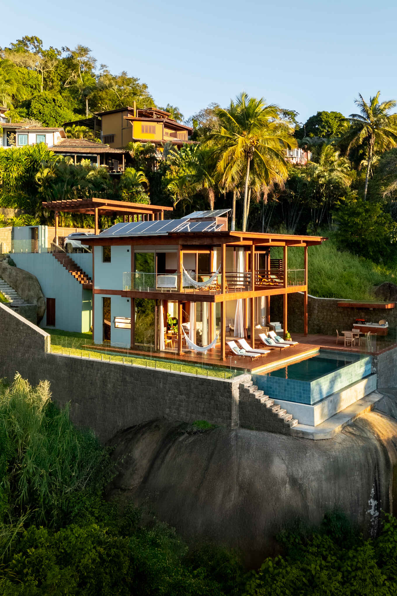 Casa Ponta da Sela in Ilhabela, Brazil, uses prefabricated timber construction, stone retaining walls, glass and timber decks to step down a steep tropical site. Existing rocks, a flat roof with solar systems and an elevated pool become part of the architecture.