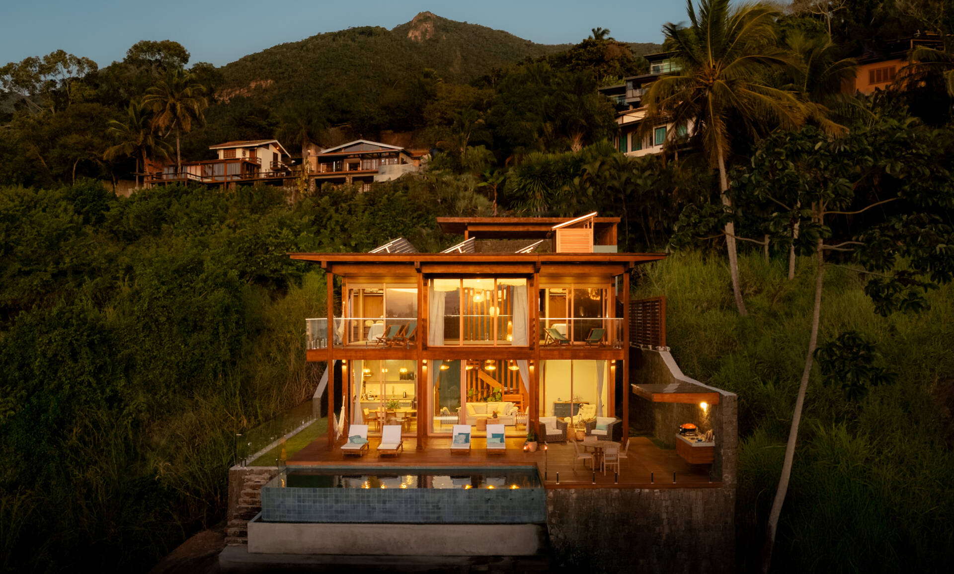 Casa Ponta da Sela in Ilhabela, Brazil, uses prefabricated timber construction, stone retaining walls, glass and timber decks to step down a steep tropical site. Existing rocks, a flat roof with solar systems and an elevated pool become part of the architecture.