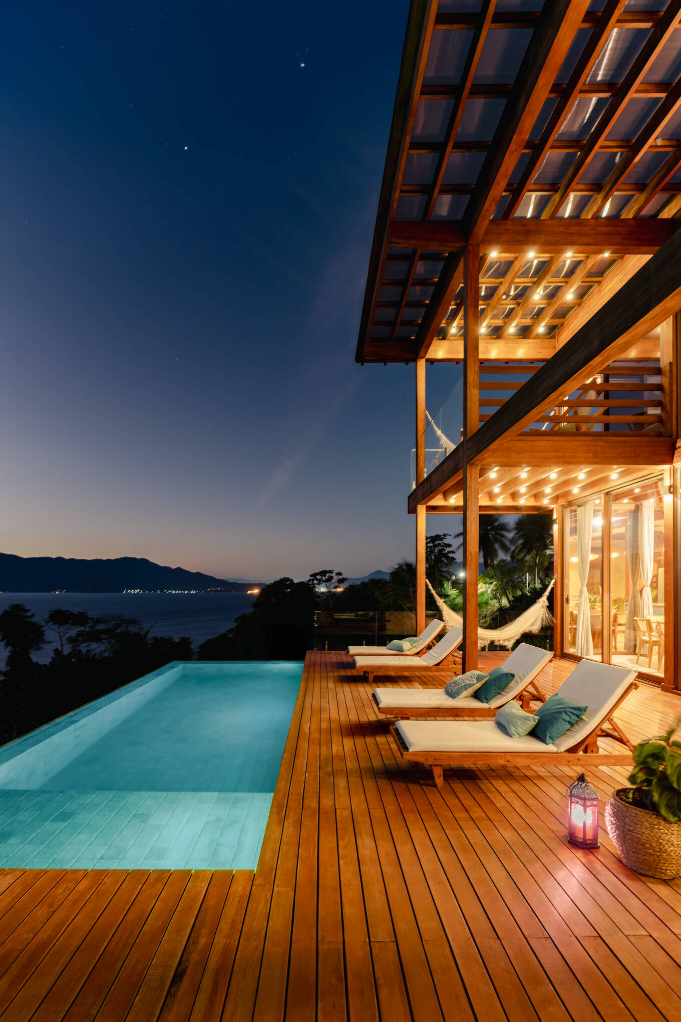 The pool and timber deck at Casa Ponta da Sela in Ilhabela extend toward sweeping water and mountain views, creating an outdoor living area with hammocks, loungers, glass balustrades and exposed timber framing.