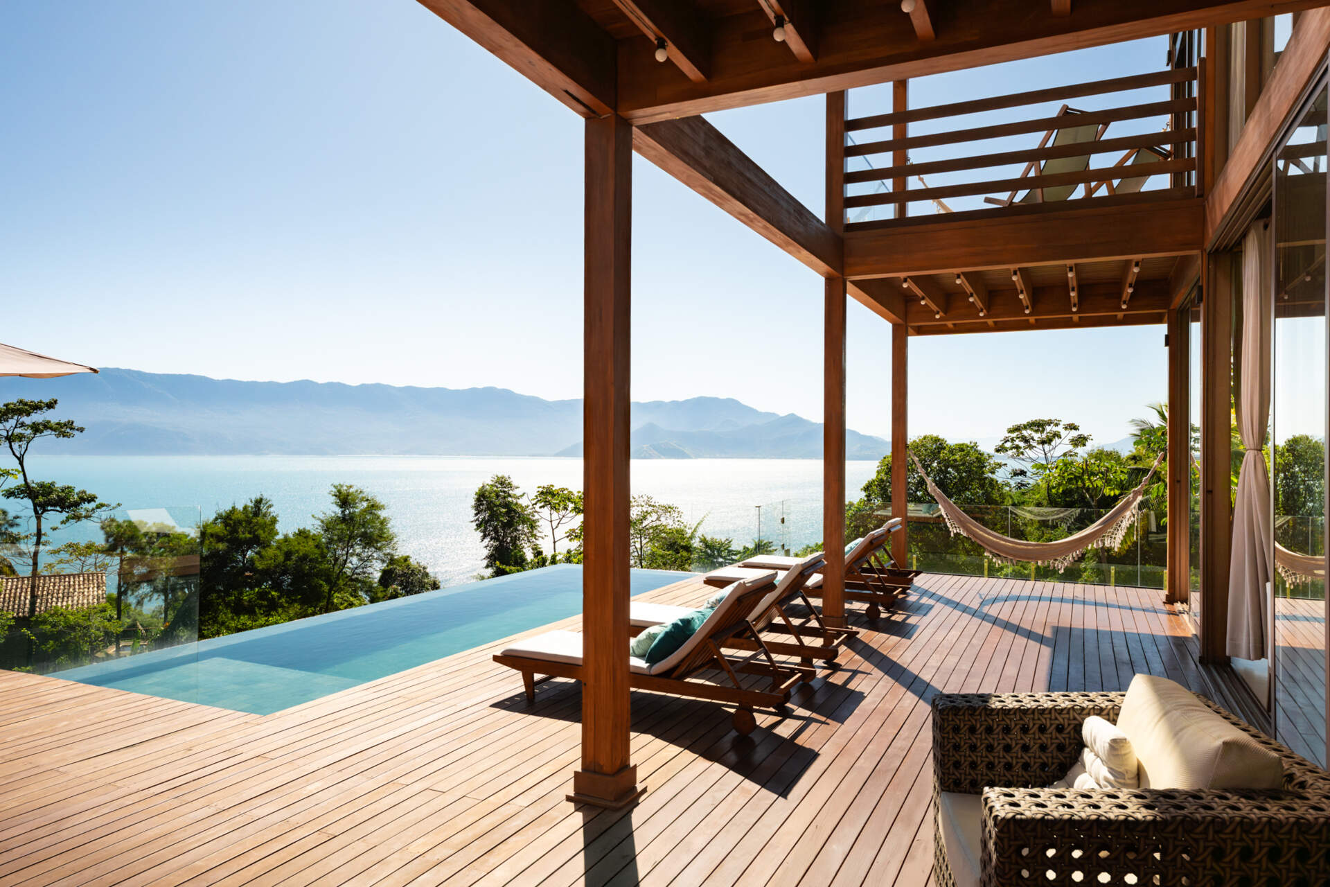 The pool and timber deck at Casa Ponta da Sela in Ilhabela extend toward sweeping water and mountain views, creating an outdoor living area with hammocks, loungers, glass balustrades and exposed timber framing.
