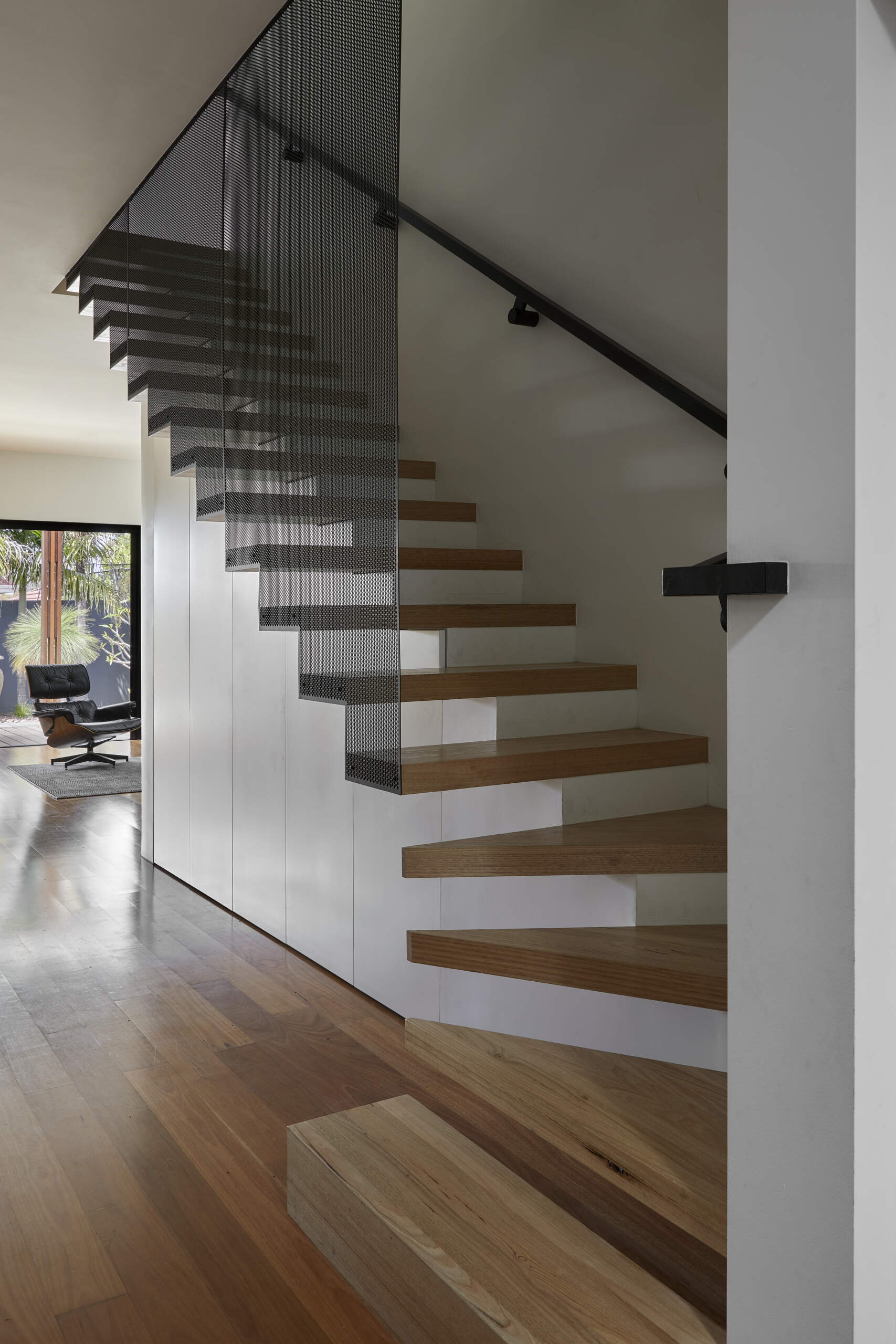 A side view of a modern staircase highlights floating timber treads, black perforated metal screening, and built-in cabinetry beneath the stairs.
