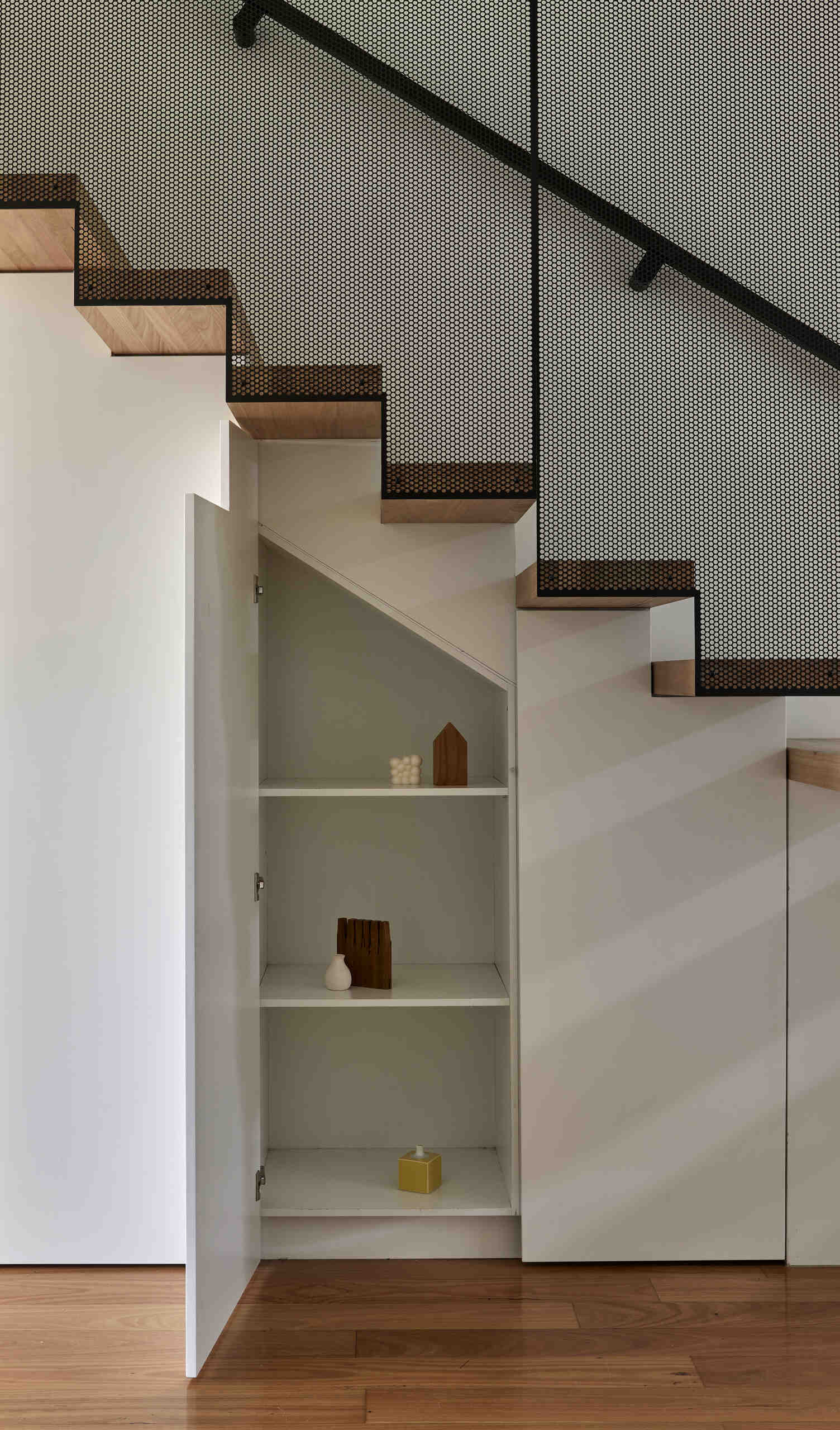 Built-in storage shelves make use of the angled space beneath a modern staircase. A white cabinet door opens to reveal shelving fitted below the timber treads and perforated metal screen.