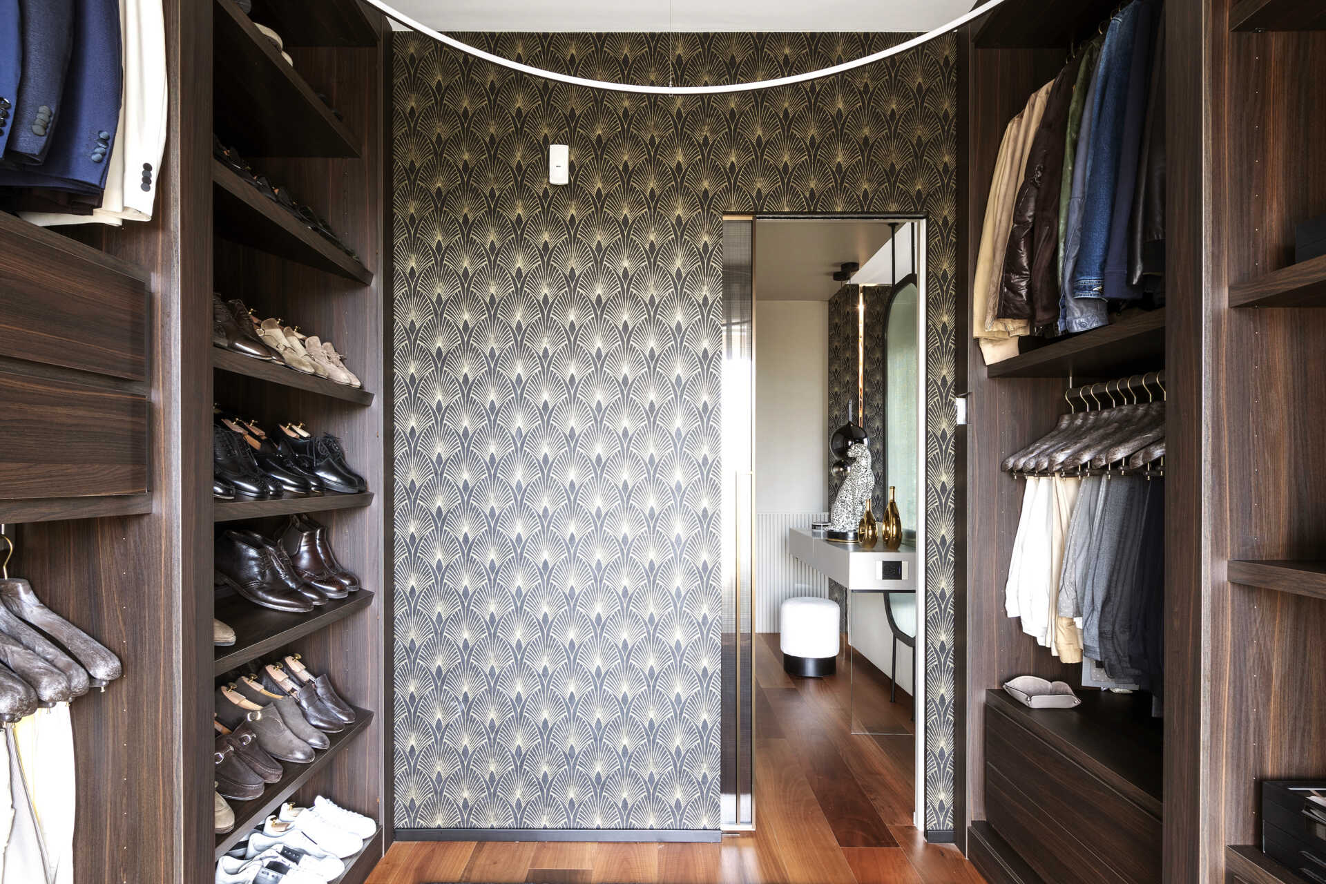A walk-in closet features dark timber cabinetry, open shoe shelves, hanging rails, built-in drawers, Art Deco wallpaper, and a curved ceiling light.