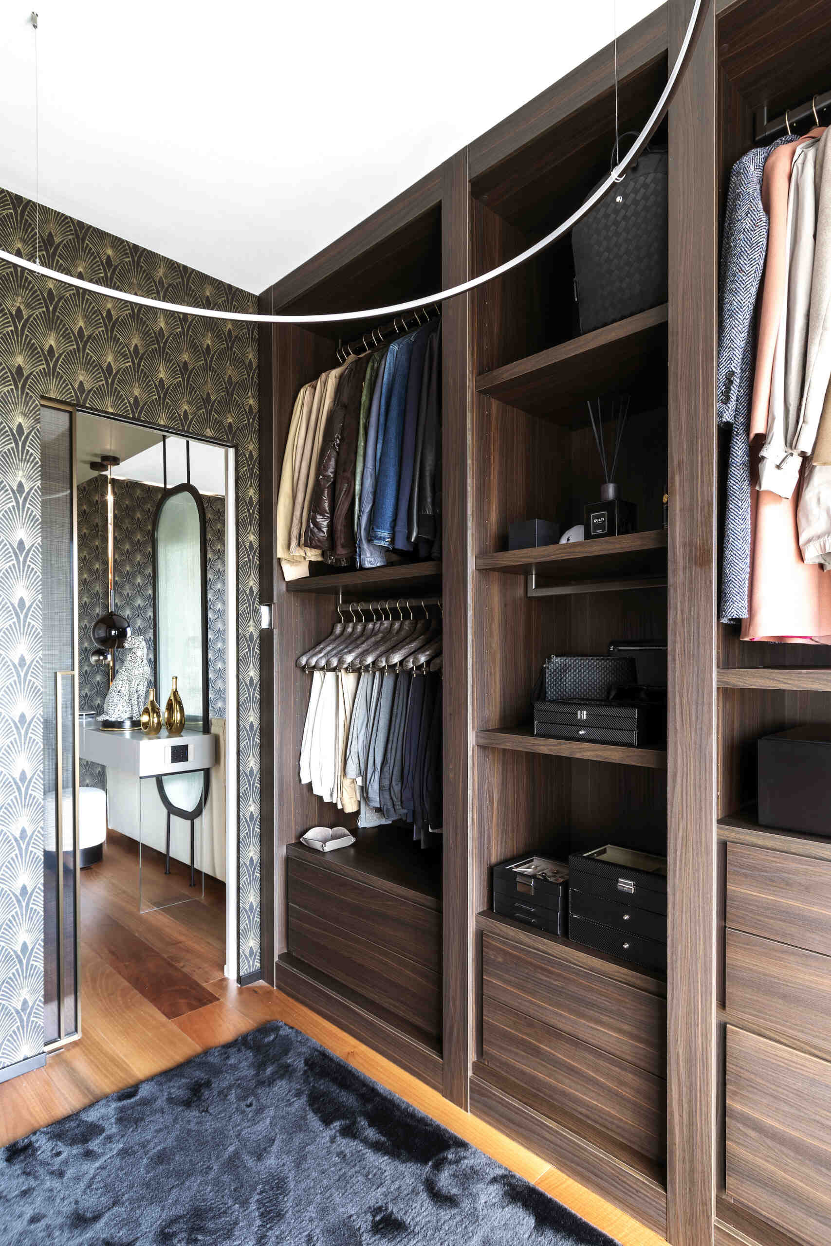 A walk-in closet features dark timber cabinetry, open shoe shelves, hanging rails, built-in drawers, Art Deco wallpaper, and a curved ceiling light.