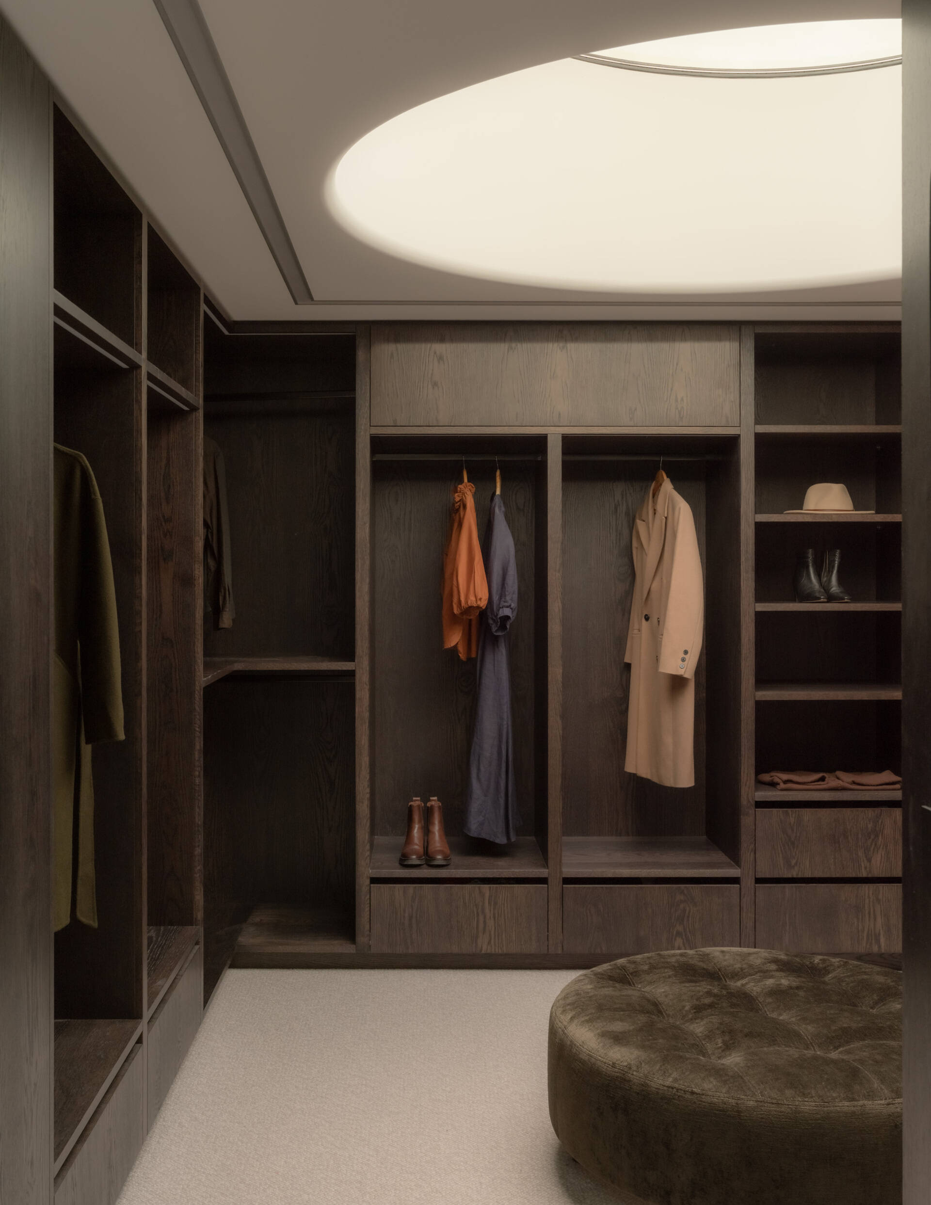 A contemporary walk-in dressing room with a large circular skylight, floor-to-ceiling dark oak wardrobes, open clothing storage, pale carpeting, and a central upholstered ottoman.
