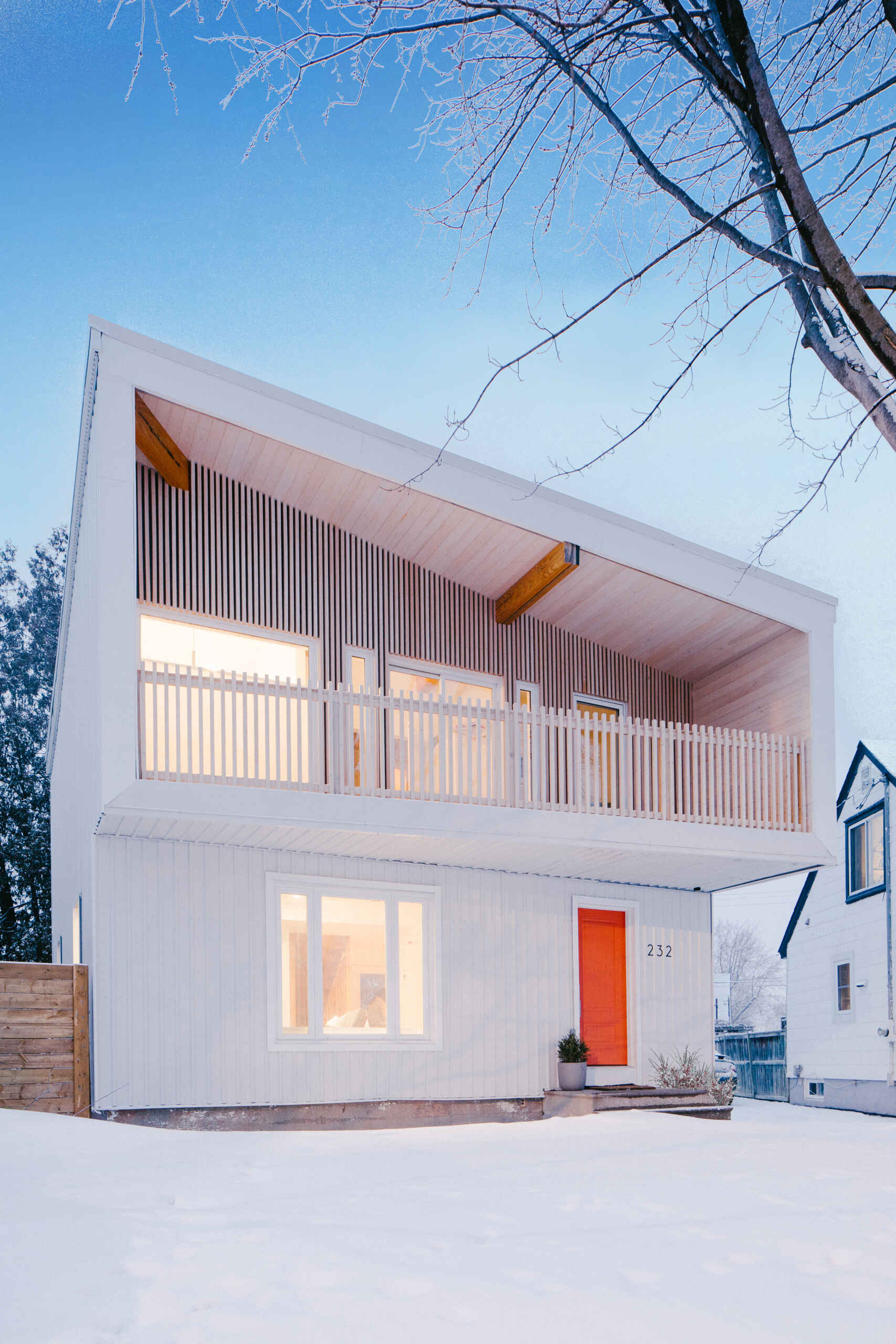 This renovated post-war house in Ottawa features white corrugated metal siding, an angular roof, a cantilevered balcony, vertical timber battens, and a bright orange front door.