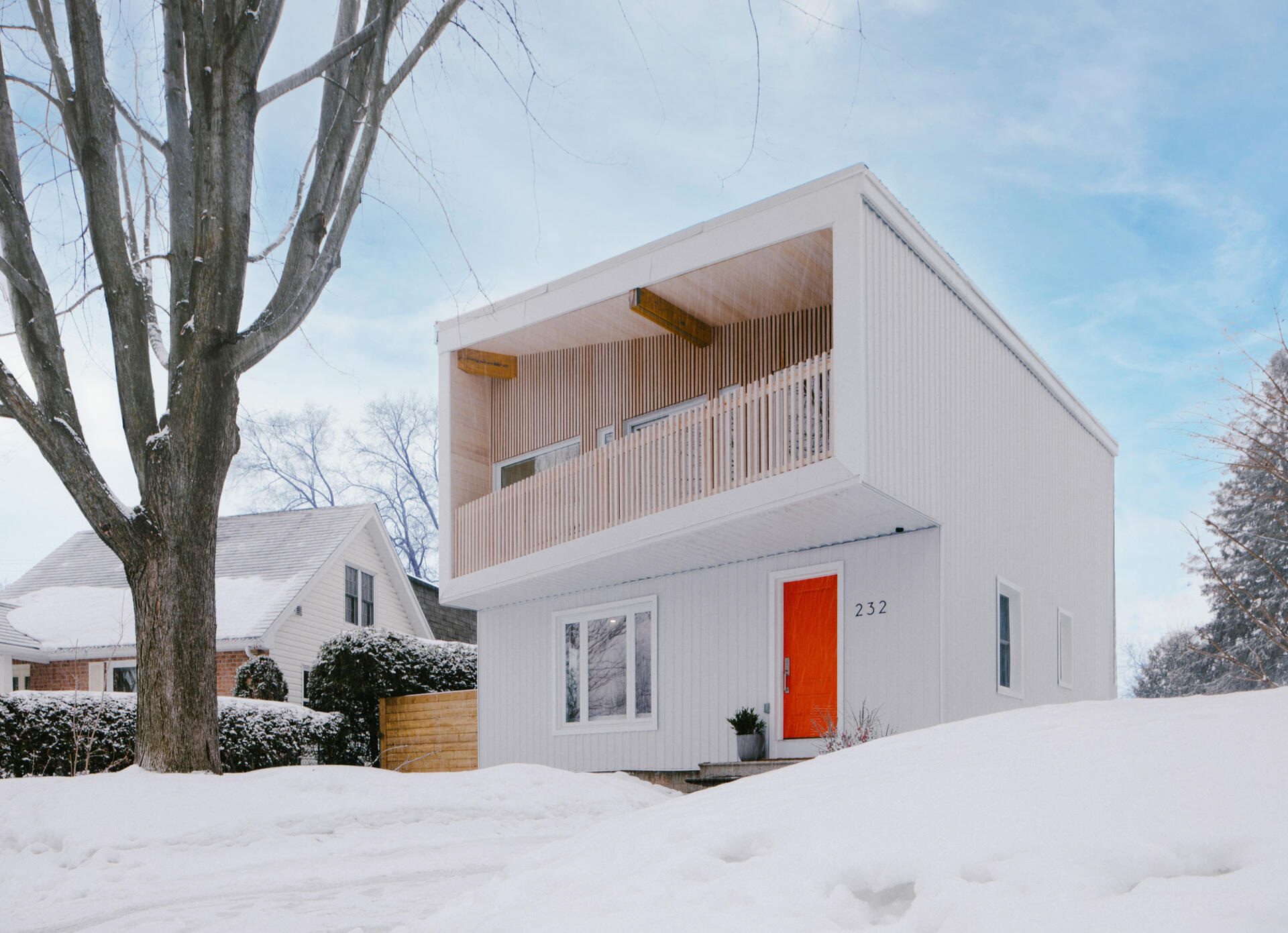 This renovated post-war house in Ottawa features white corrugated metal siding, an angular roof, a cantilevered balcony, vertical timber battens, and a bright orange front door.