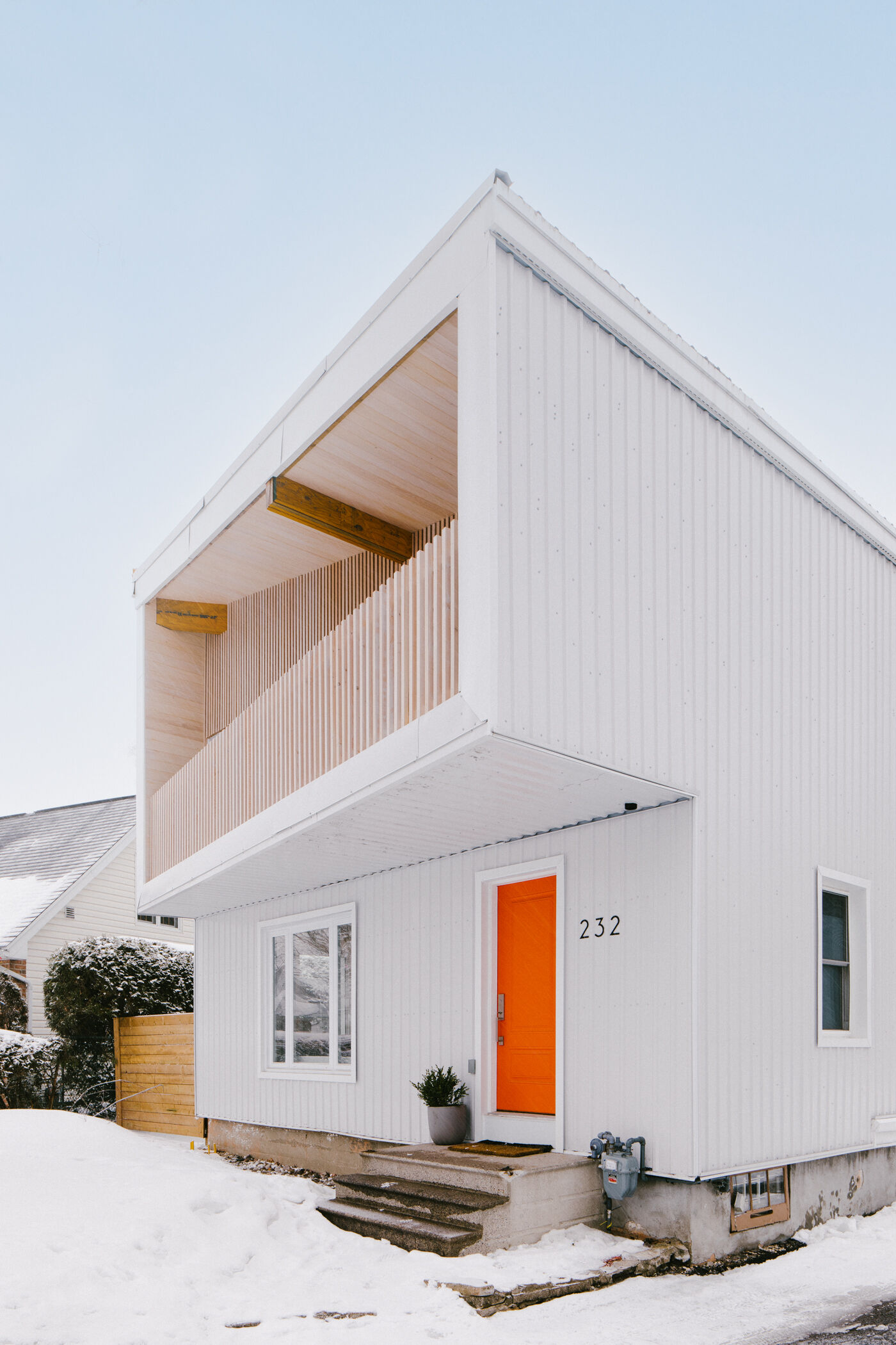 This renovated post-war house in Ottawa features white corrugated metal siding, an angular roof, a cantilevered balcony, vertical timber battens, and a bright orange front door.