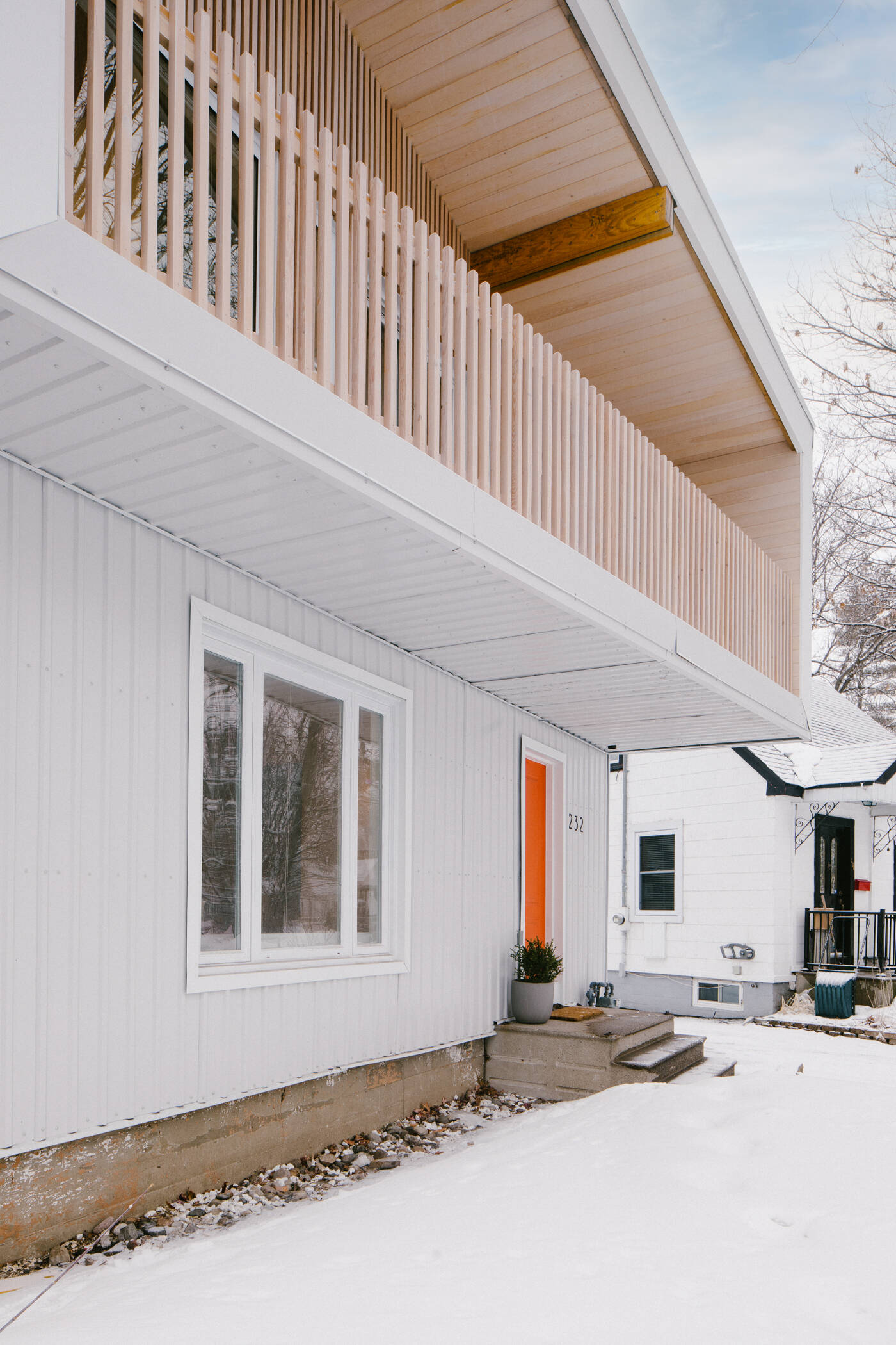 This renovated post-war house in Ottawa features white corrugated metal siding, an angular roof, a cantilevered balcony, vertical timber battens, and a bright orange front door.