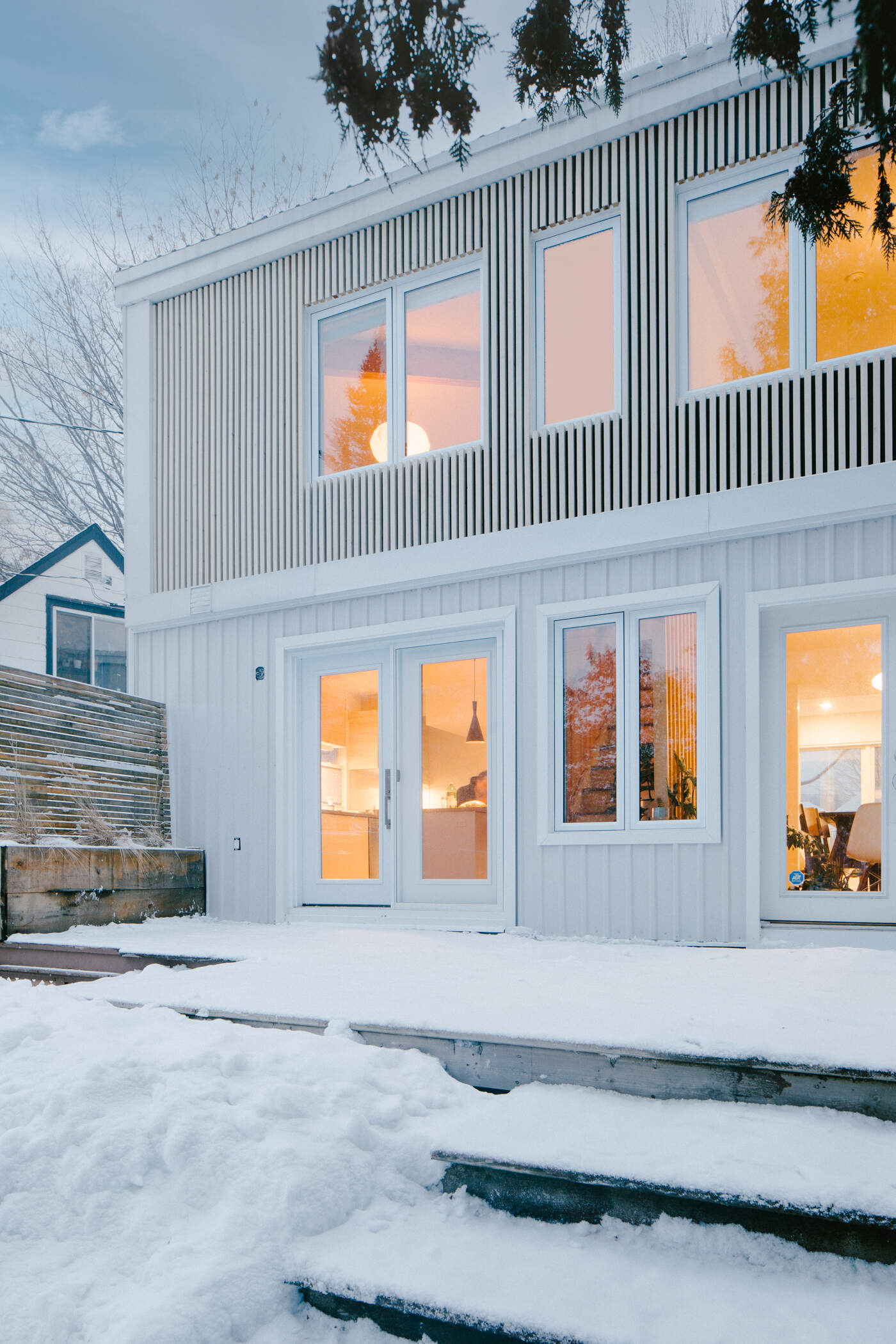 Explore the rear facade of this modern Ottawa home renovation, where large glass doors, white corrugated siding, and vertical timber screens create a bright and welcoming design.
