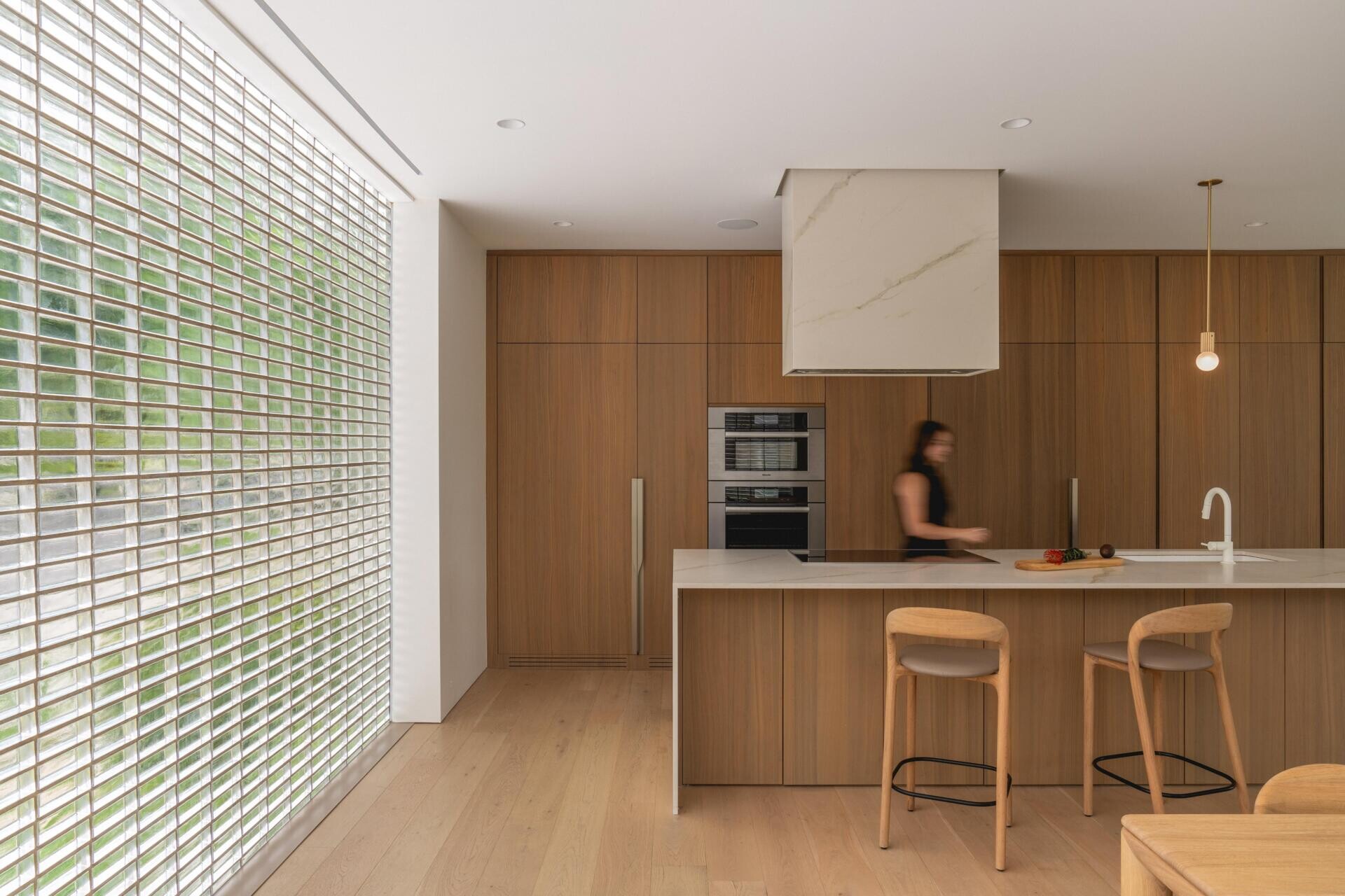 Explore a modern walnut kitchen in The Lens by MU Architecture. Full-height wood cabinetry, a minimalist porcelain island, sculptural range hood, brass pendant lighting, and wooden bar stools define this Montreal home's contemporary design.