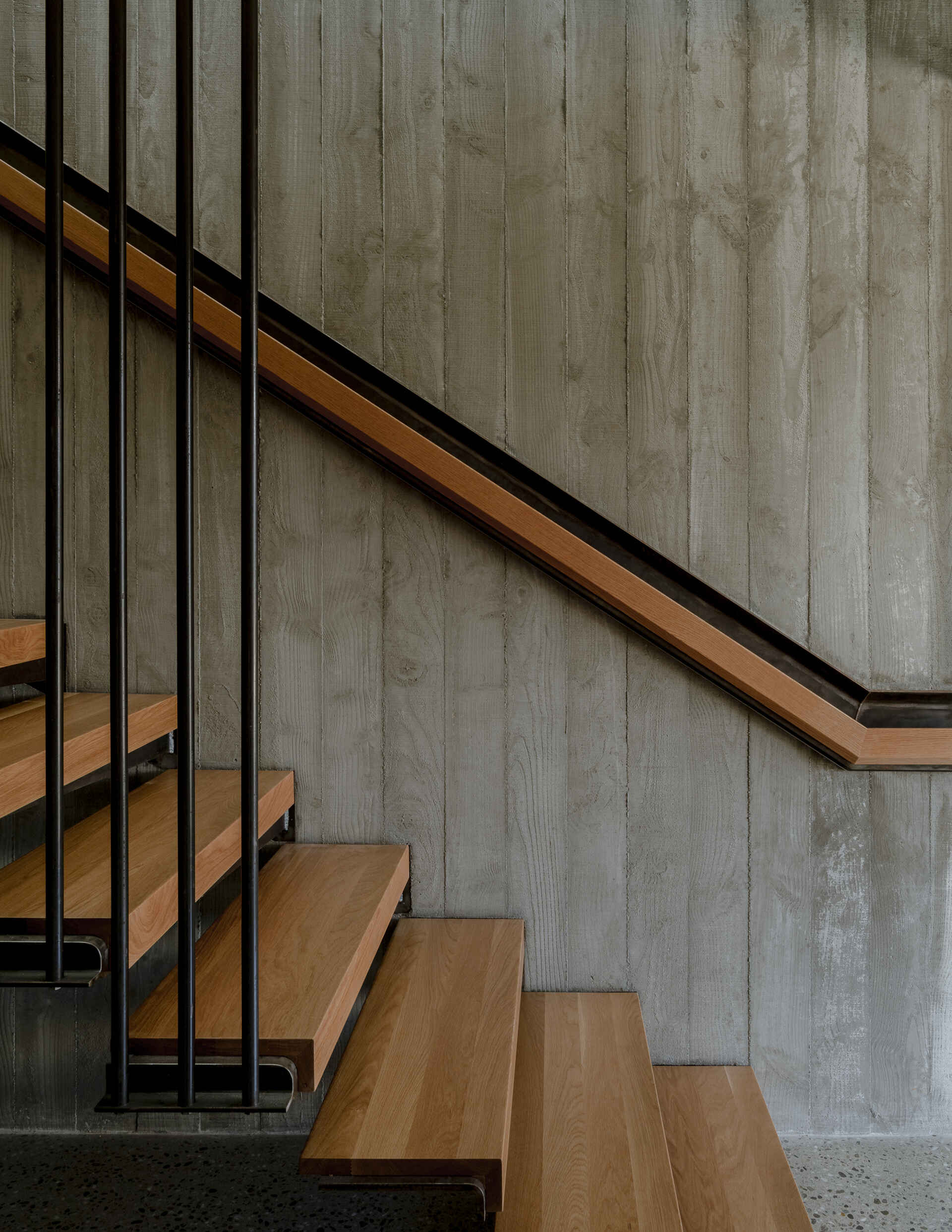 Modern staircase with open wood steps, slim black metal railings, and textured concrete walls. Natural sunlight enters through an overhead opening, illuminating the narrow stairwell and glass door leading to an outdoor deck.