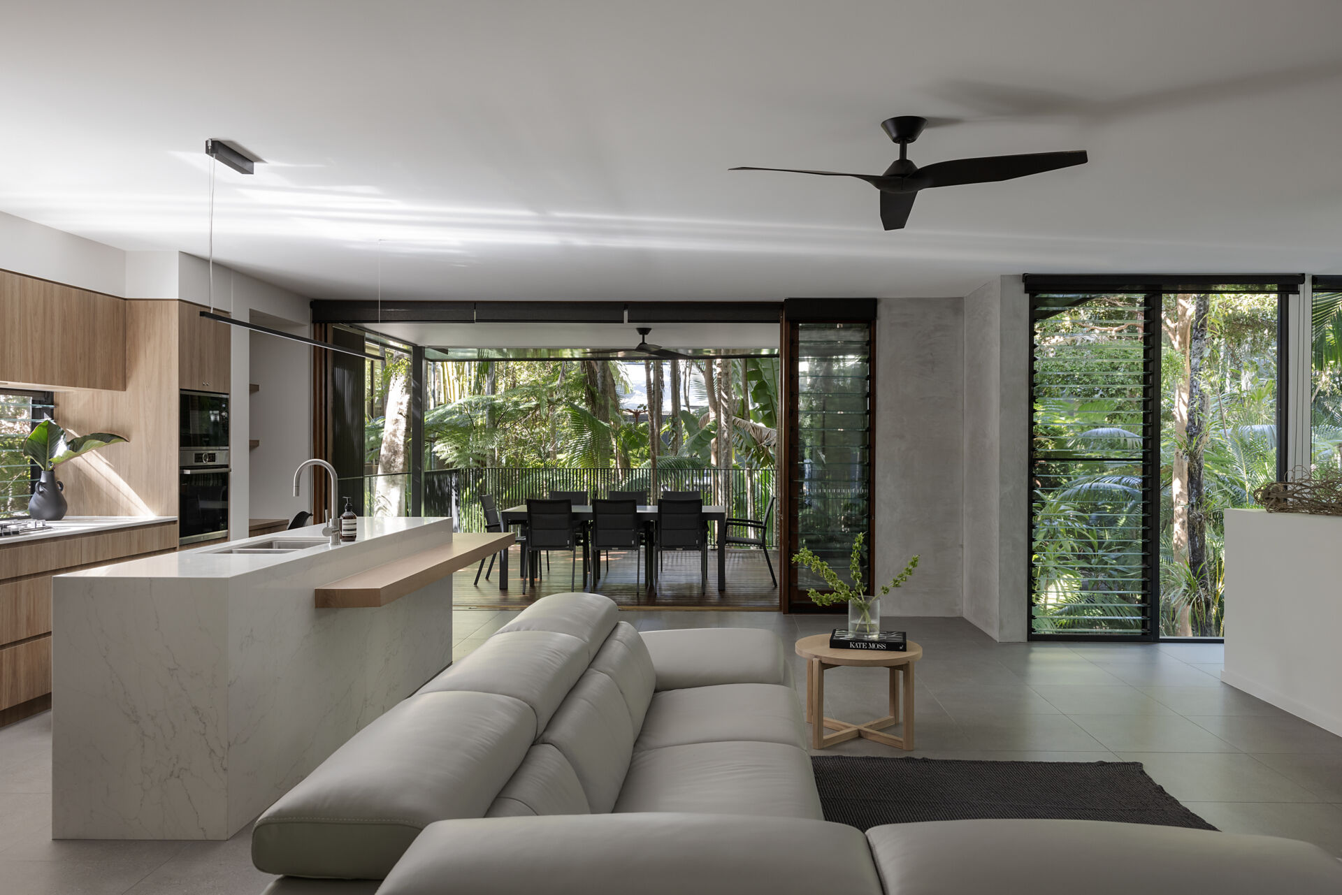 Take a look inside the living room of Raintree in Noosa Heads, Australia. A white sectional sofa, natural timber furniture, grey tiled floors, and floor-to-ceiling windows create a modern coastal interior with lush rainforest views.