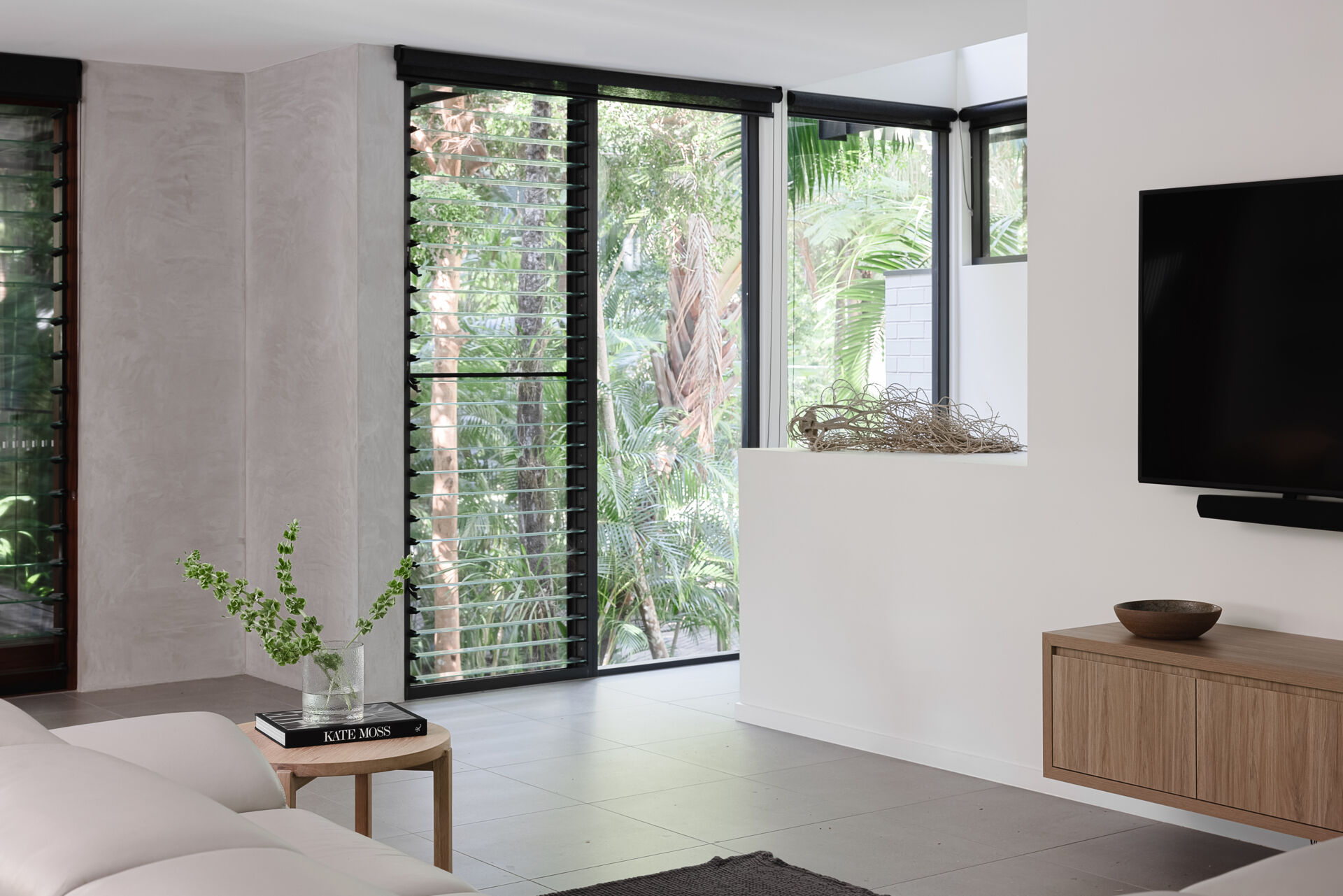 Take a look inside the living room of Raintree in Noosa Heads, Australia. A white sectional sofa, natural timber furniture, grey tiled floors, and floor-to-ceiling windows create a modern coastal interior with lush rainforest views.