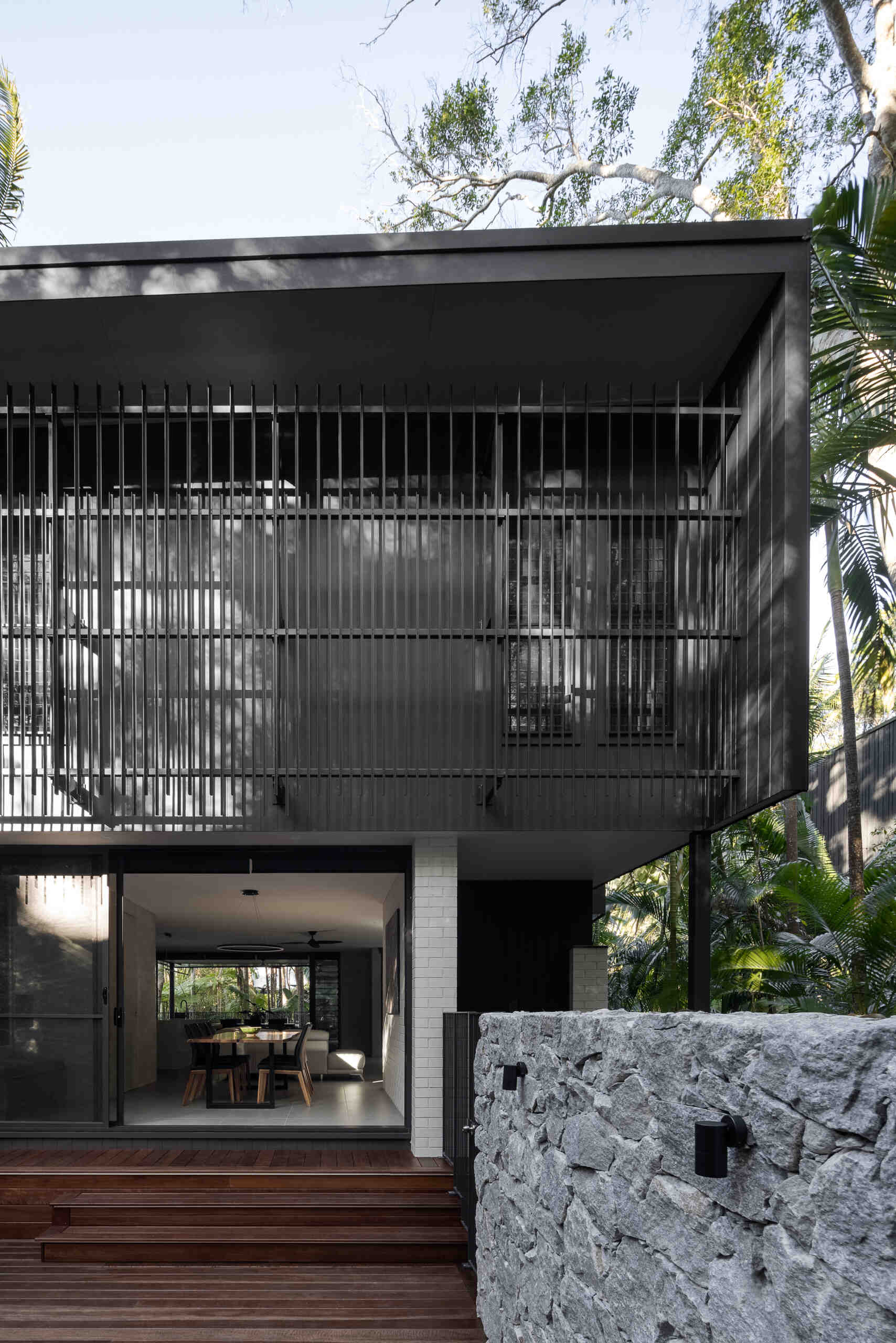 Explore the private patio at Raintree in Noosa Heads, designed by Void Building Design. A timber deck, built-in outdoor bench, natural stone walls, concrete stepping stones, and lush tropical landscaping create a secluded outdoor living space.