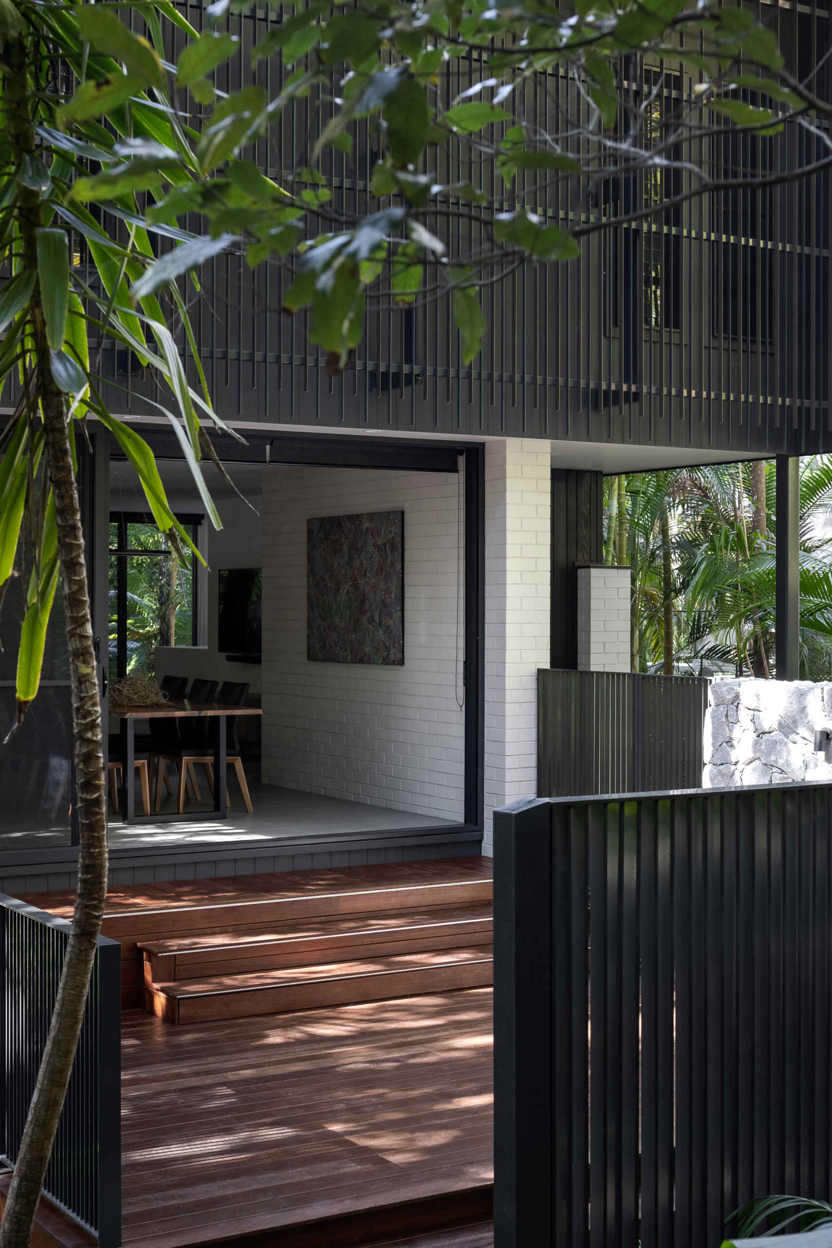 Explore the private patio at Raintree in Noosa Heads, designed by Void Building Design. A timber deck, built-in outdoor bench, natural stone walls, concrete stepping stones, and lush tropical landscaping create a secluded outdoor living space.