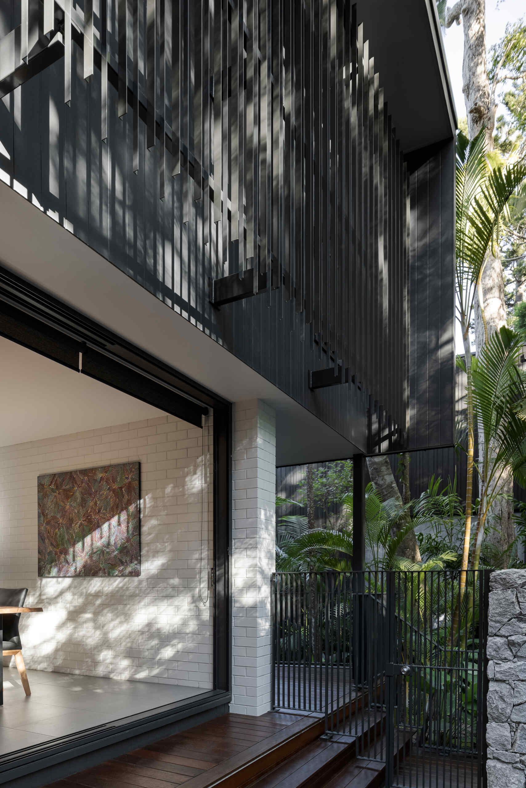 Explore the private patio at Raintree in Noosa Heads, designed by Void Building Design. A timber deck, built-in outdoor bench, natural stone walls, concrete stepping stones, and lush tropical landscaping create a secluded outdoor living space.