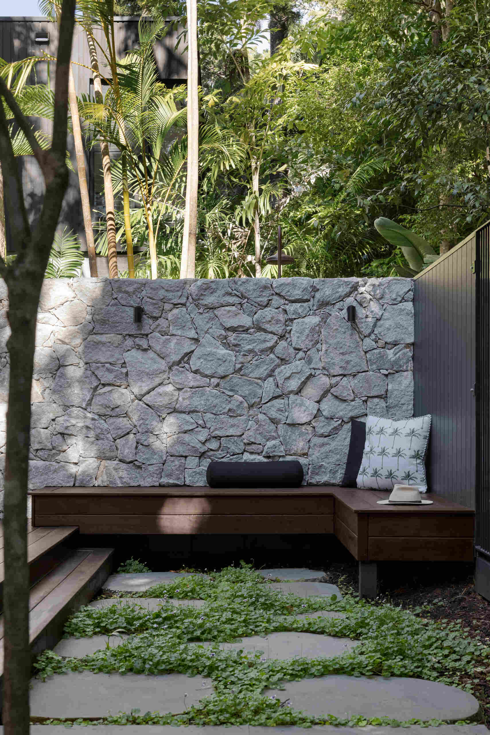 Explore the private patio at Raintree in Noosa Heads, designed by Void Building Design. A timber deck, built-in outdoor bench, natural stone walls, concrete stepping stones, and lush tropical landscaping create a secluded outdoor living space.