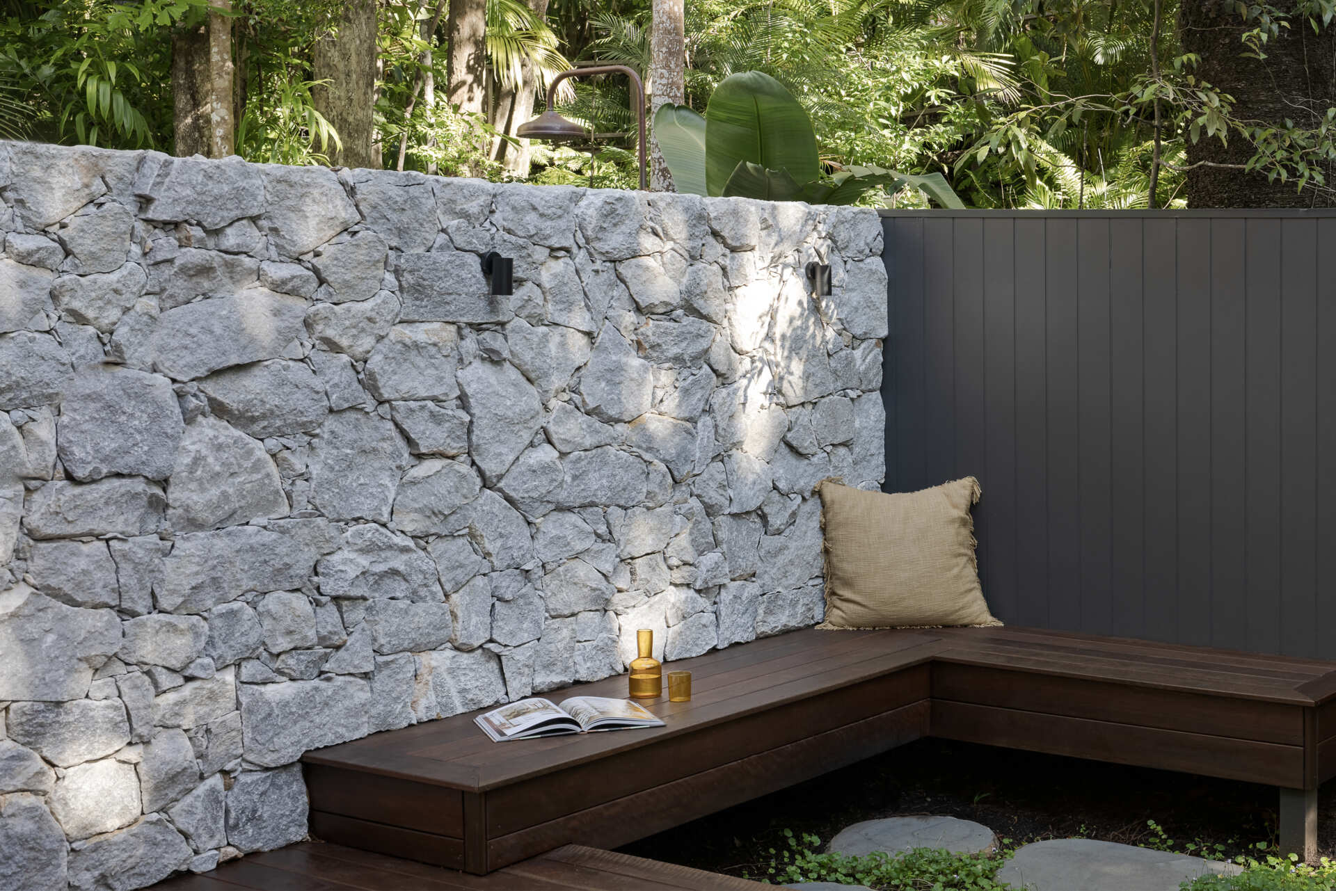 Explore the private patio at Raintree in Noosa Heads, designed by Void Building Design. A timber deck, built-in outdoor bench, natural stone walls, concrete stepping stones, and lush tropical landscaping create a secluded outdoor living space.