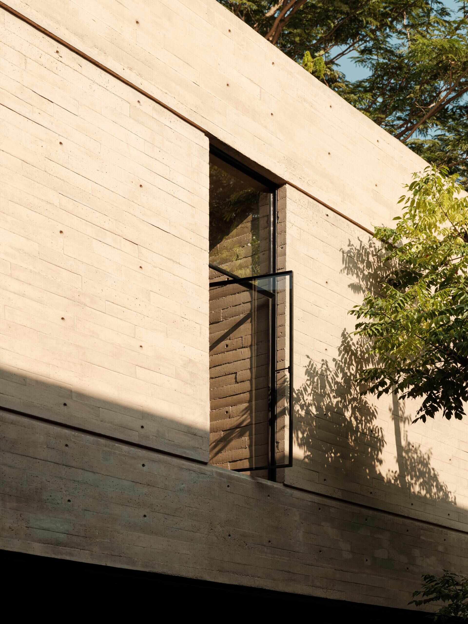 A tall pivoting glass window in a modern Mexican home features a slim black steel frame, exposed concrete walls, and views of a leafy courtyard. A unique architectural window design that brings light and ventilation indoors.
