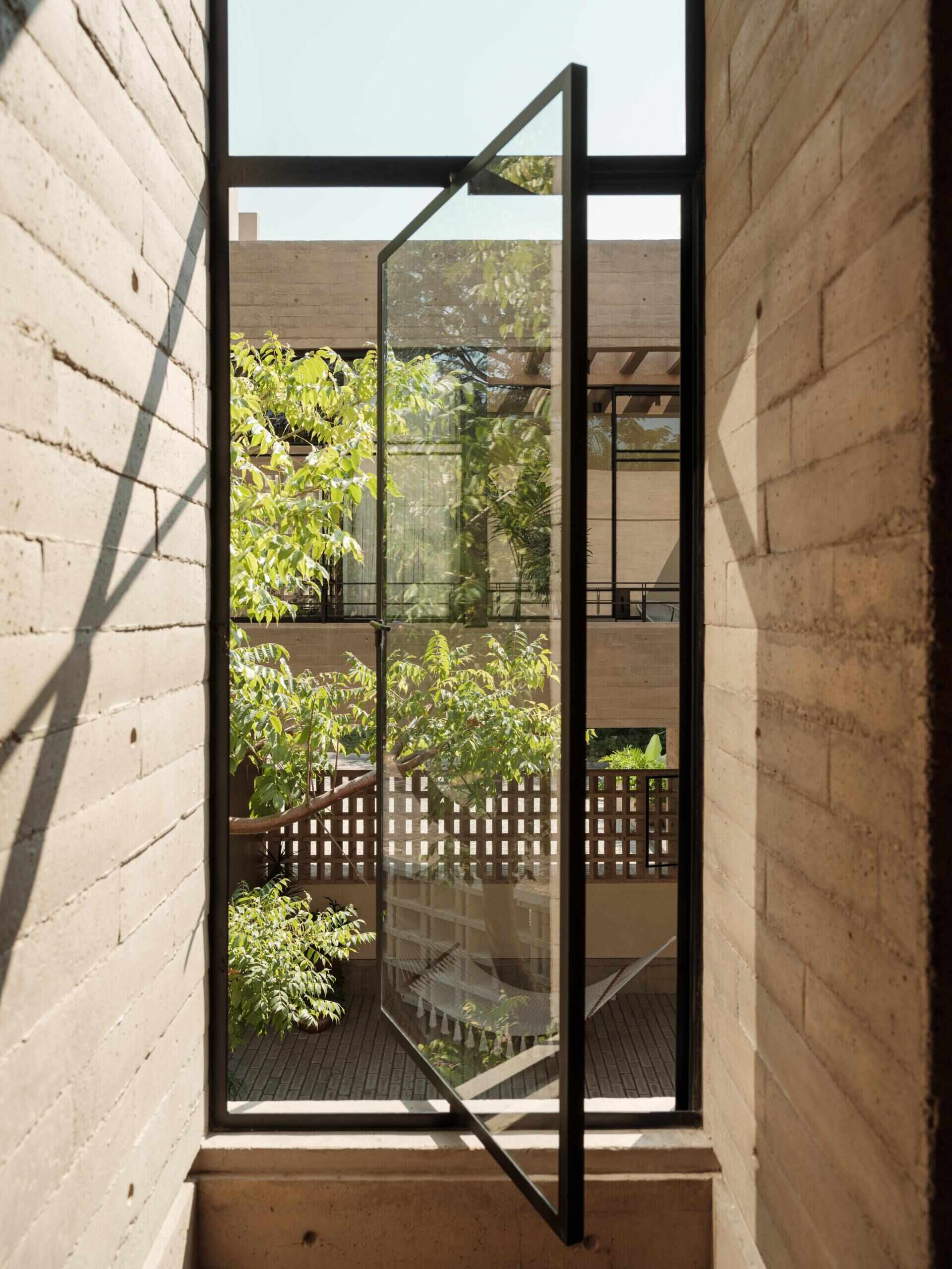 A tall pivoting glass window in a modern Mexican home features a slim black steel frame, exposed concrete walls, and views of a leafy courtyard. A unique architectural window design that brings light and ventilation indoors.