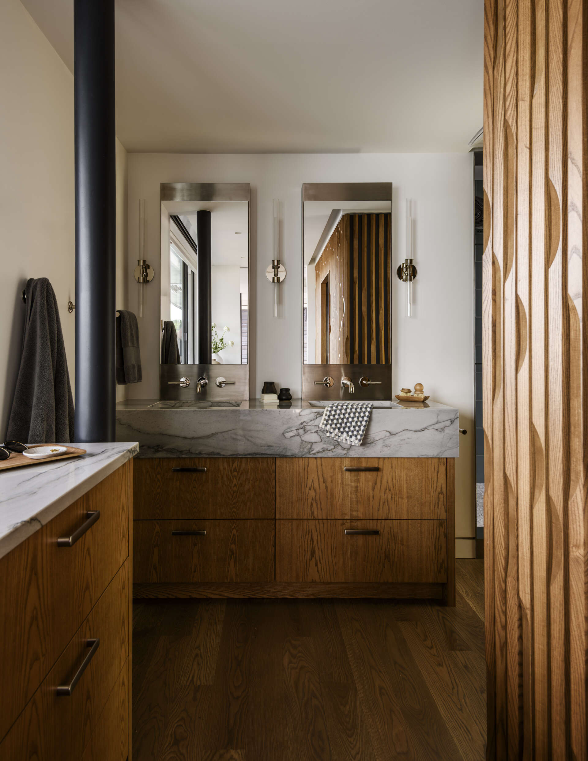 Featuring a double vanity with warm wood cabinets, gray-veined countertops, a blue-tiled walk-in shower, pebble mosaic flooring, and a large frosted window, this modern bathroom combines natural textures with coastal-inspired colors.
