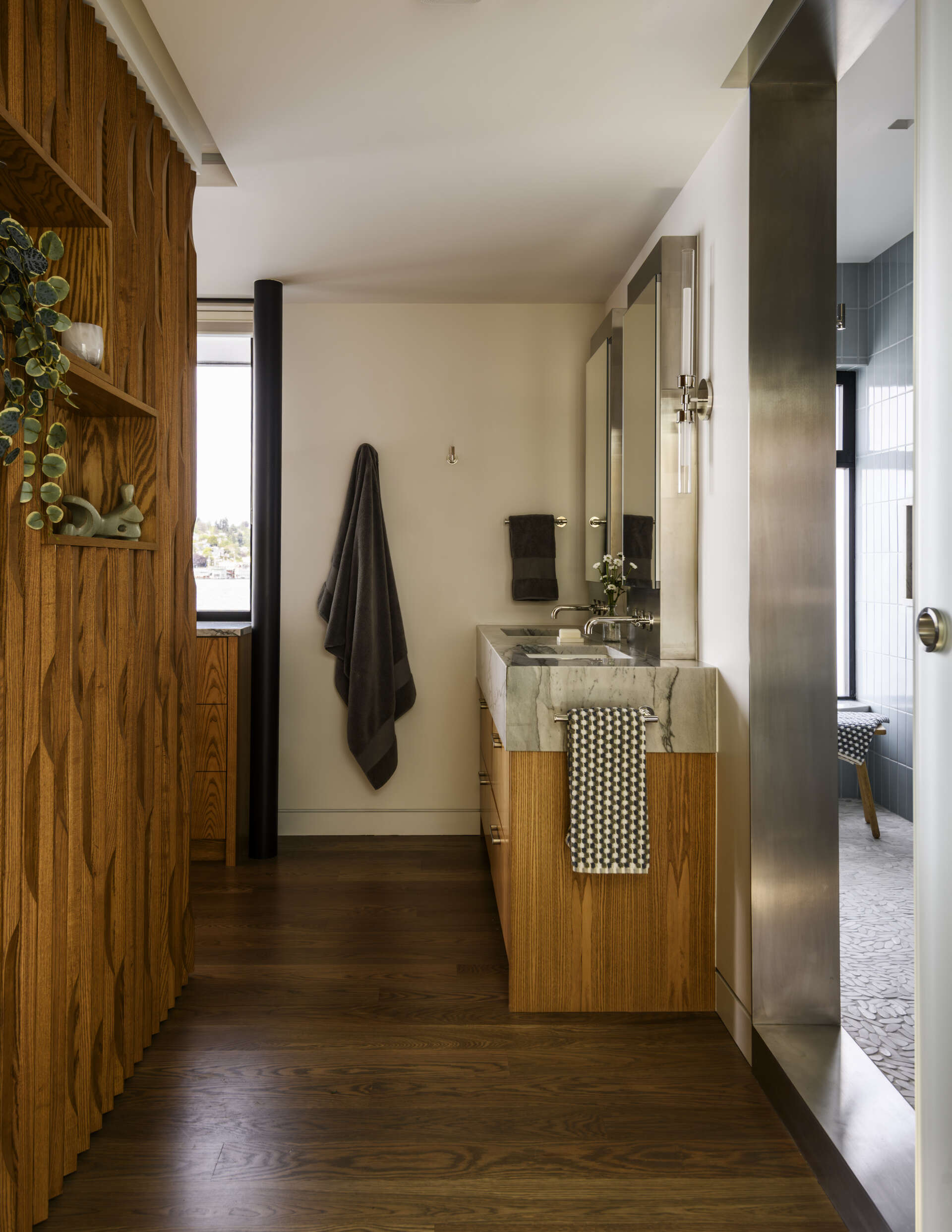 Featuring a double vanity with warm wood cabinets, gray-veined countertops, a blue-tiled walk-in shower, pebble mosaic flooring, and a large frosted window, this modern bathroom combines natural textures with coastal-inspired colors.