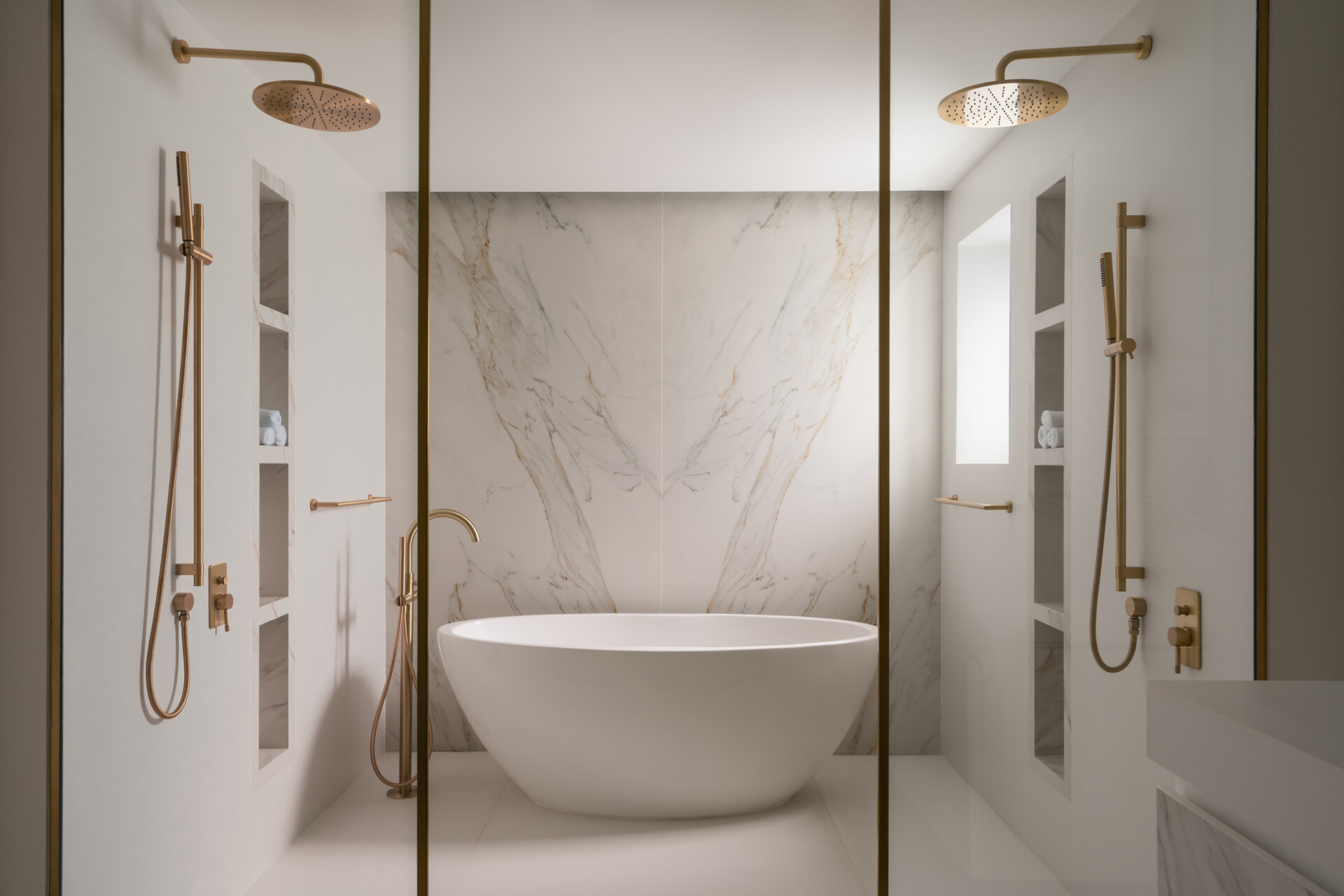 Explore the luxurious primary bathroom at The White Arcade in Dubai. Featuring a freestanding oval bathtub, bookmatched marble feature wall, dual rainfall showers, brass bathroom fixtures, and built-in marble niches, this elegant bathroom was designed by D’Ora Tokai Designs.