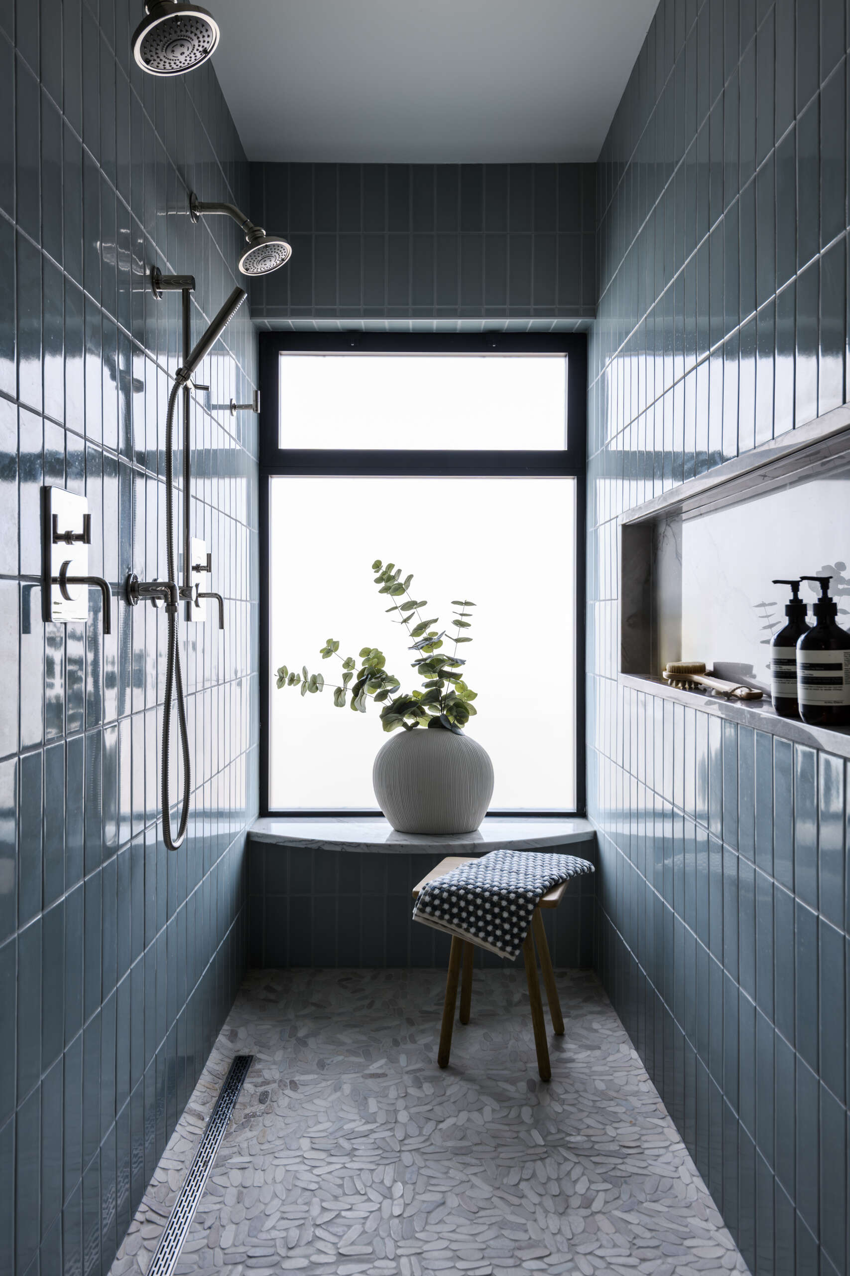 Featuring a double vanity with warm wood cabinets, gray-veined countertops, a blue-tiled walk-in shower, pebble mosaic flooring, and a large frosted window, this modern bathroom combines natural textures with coastal-inspired colors.