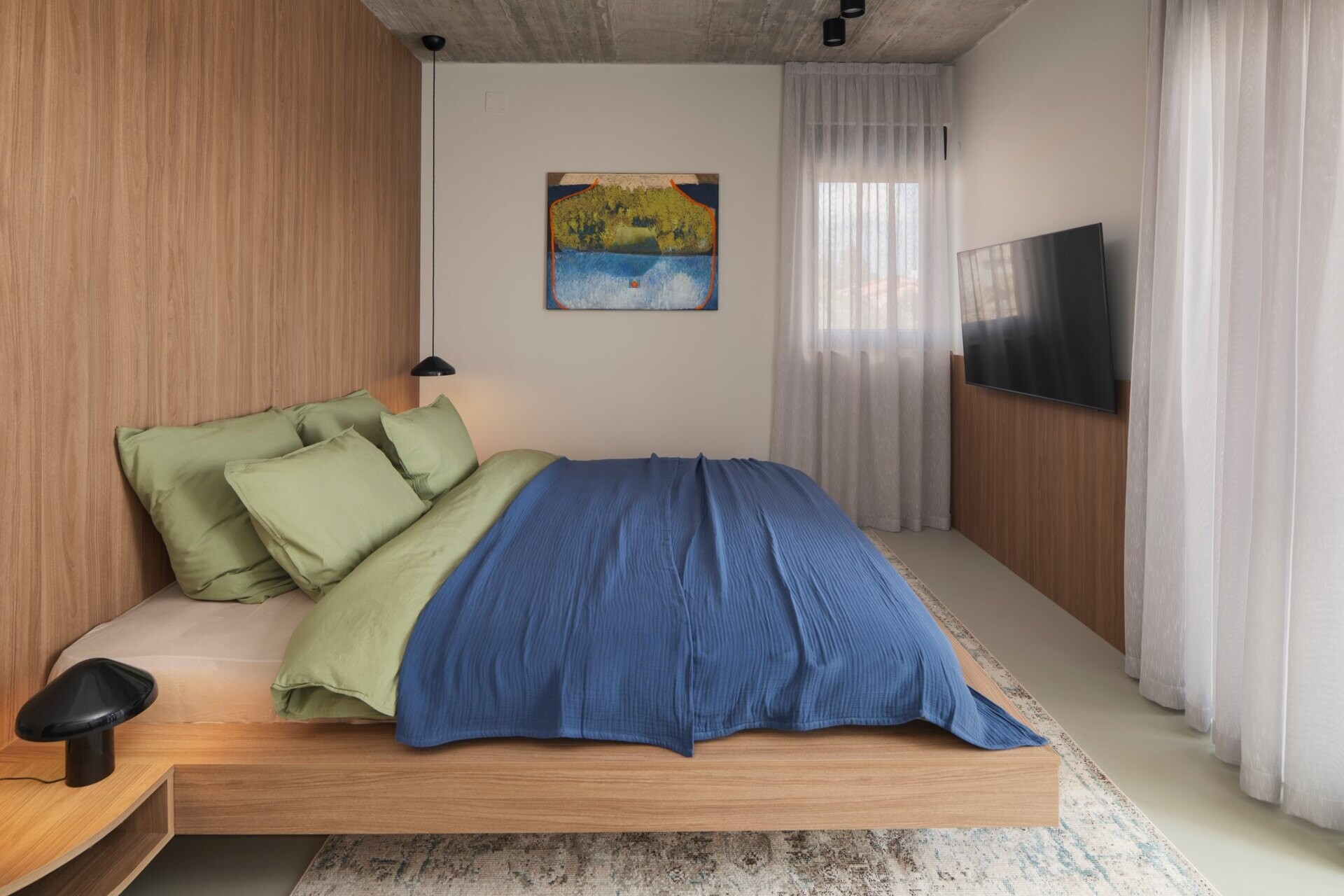 Tour the primary bedroom suite at Arc House in Croatia, featuring a timber platform bed, blue and green bedding, built-in wardrobes, a private sitting area, open wooden shelving and access to a covered terrace.