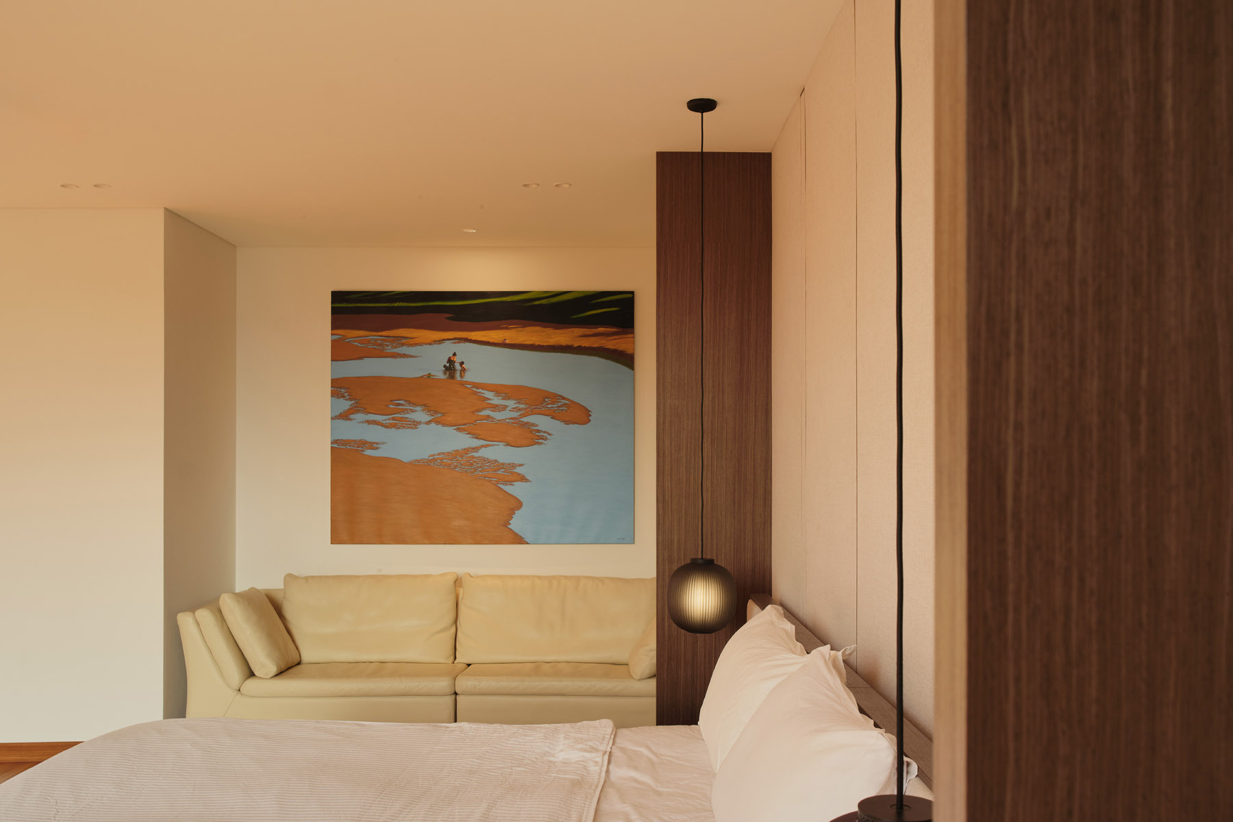 Take a look inside the primary bedroom of JS-House, featuring warm timber floors, a fabric headboard wall, floating bedside furniture, pendant lighting, and a private sitting area.