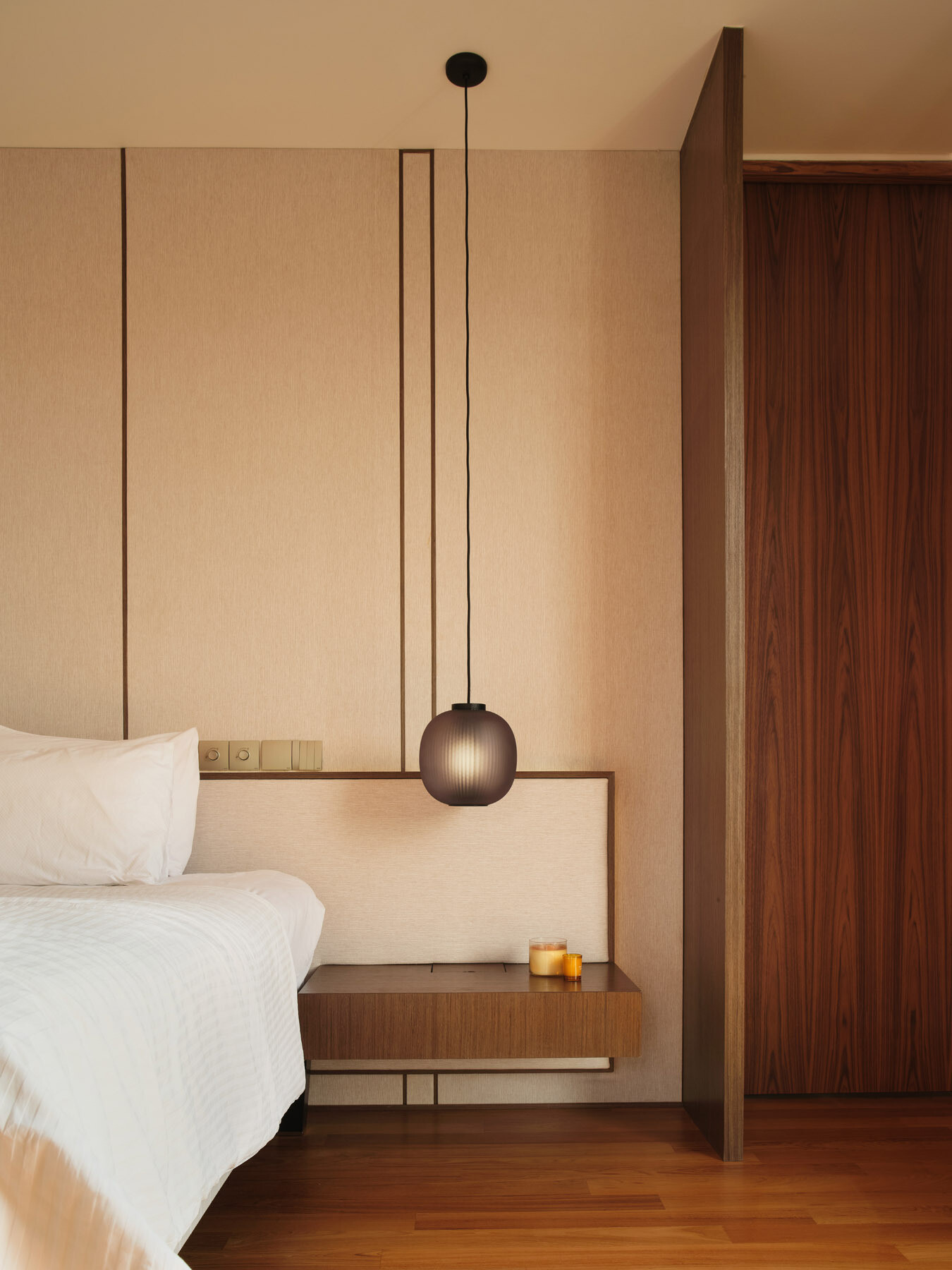 Take a look inside the primary bedroom of JS-House, featuring warm timber floors, a fabric headboard wall, floating bedside furniture, pendant lighting, and a private sitting area.