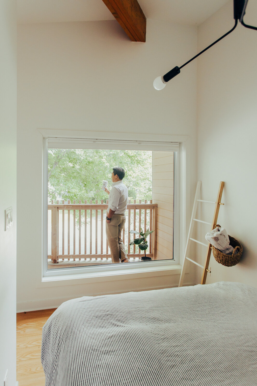 This primary bedroom takes advantage of the house's new roof profile and its position overlooking the mature maple tree in the front yard. A large window looks out toward the sheltered balcony, bringing greenery and daylight directly into the bedroom. The relatively compact space is arranged to accommodate both sleeping and working, while white walls, wooden floors, and simple furniture help maintain an uncluttered appearance.