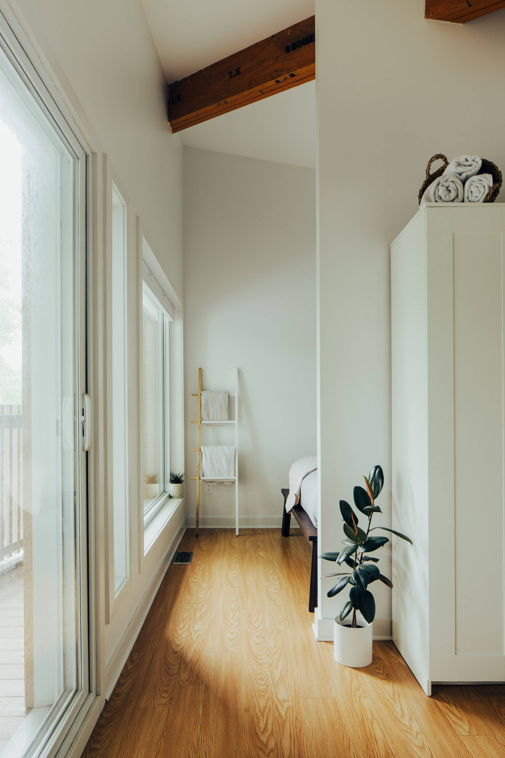 This primary bedroom takes advantage of the house's new roof profile and its position overlooking the mature maple tree in the front yard. A large window looks out toward the sheltered balcony, bringing greenery and daylight directly into the bedroom. The relatively compact space is arranged to accommodate both sleeping and working, while white walls, wooden floors, and simple furniture help maintain an uncluttered appearance.
