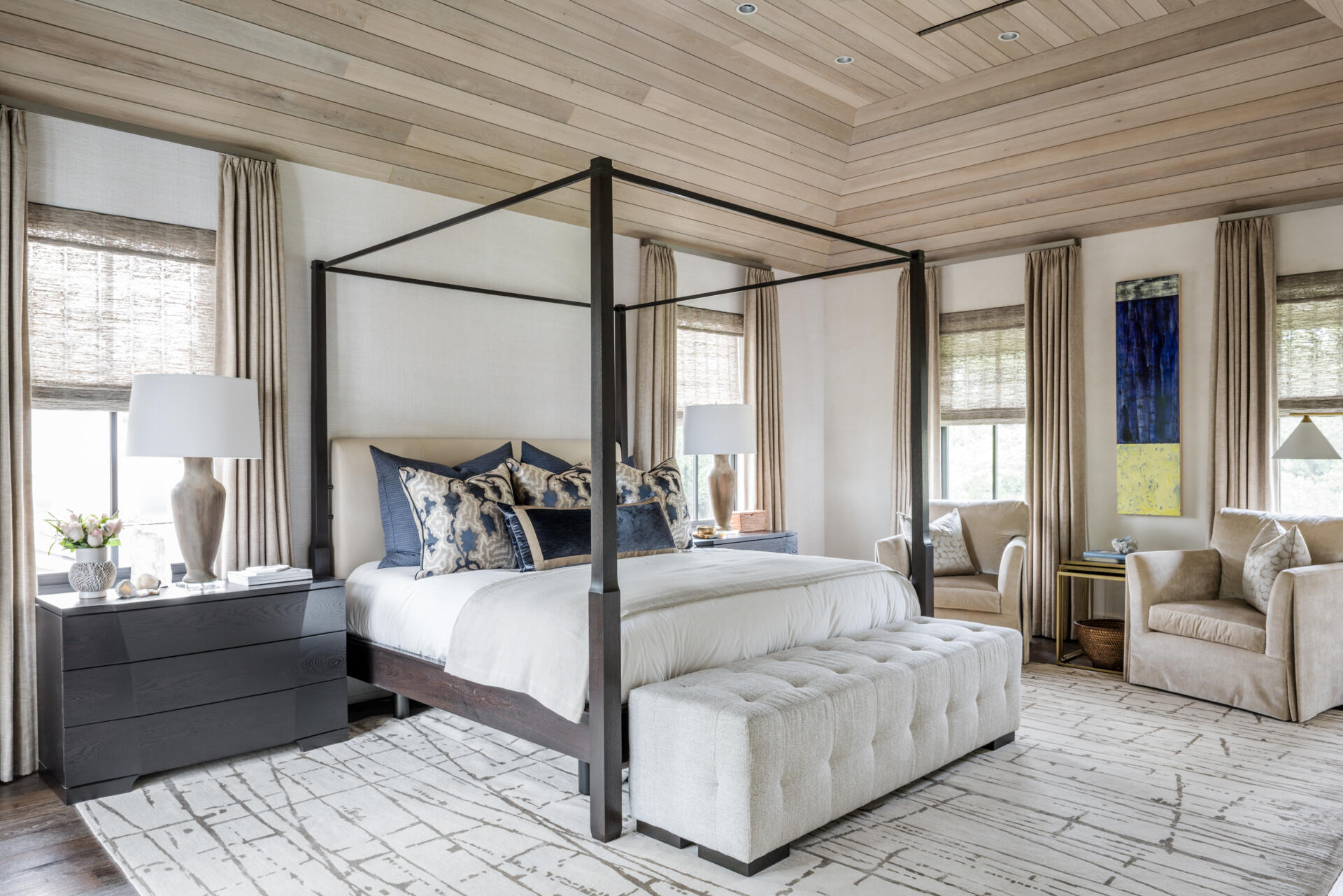 A contemporary primary bedroom with a dark four-poster bed, pale wood-paneled ceiling, blue-and-white bedding, tufted bench, patterned rug, and comfortable window-side seating.