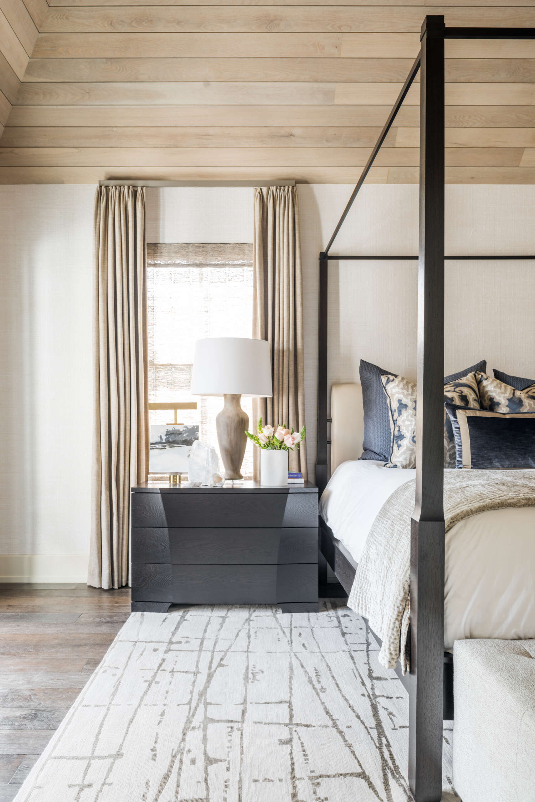 A contemporary primary bedroom with a dark four-poster bed, pale wood-paneled ceiling, blue-and-white bedding, tufted bench, patterned rug, and comfortable window-side seating.
