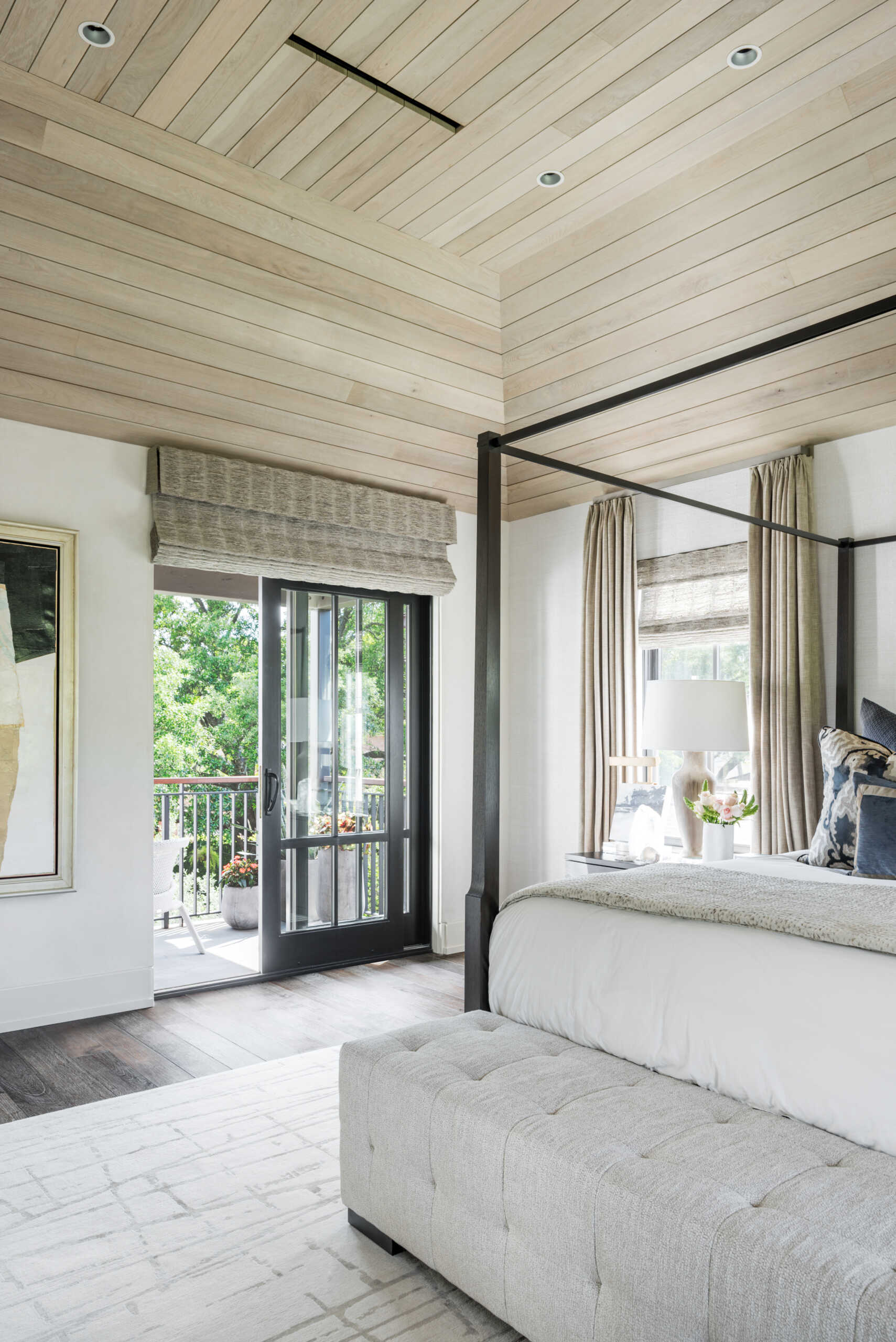 A contemporary primary bedroom with a dark four-poster bed, pale wood-paneled ceiling, blue-and-white bedding, tufted bench, patterned rug, and comfortable window-side seating.